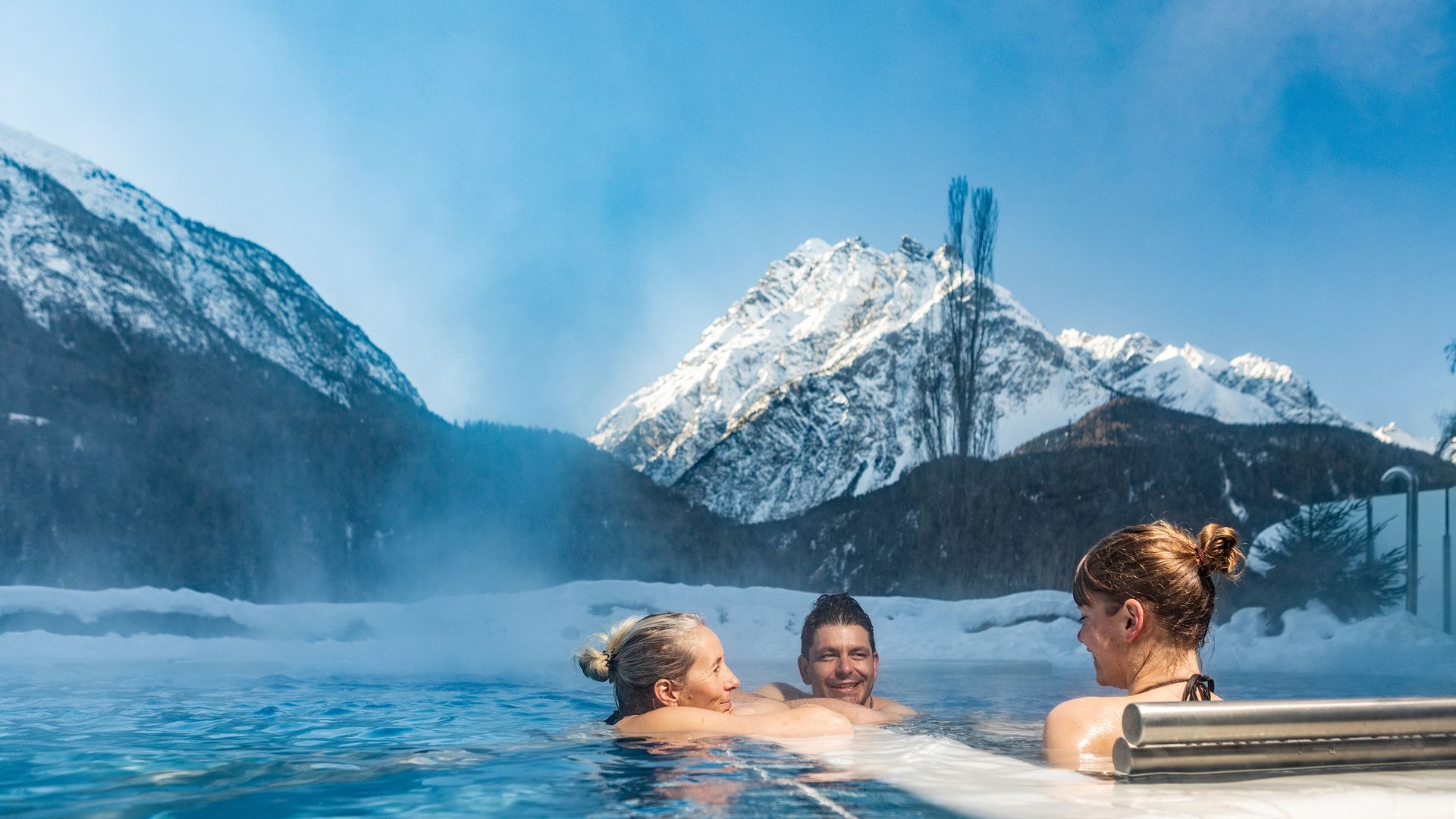 4 persone si godono il calore della piscina all'aperto e si sdraiano a bordo vasca. Il sole splende sulle cime delle montagne