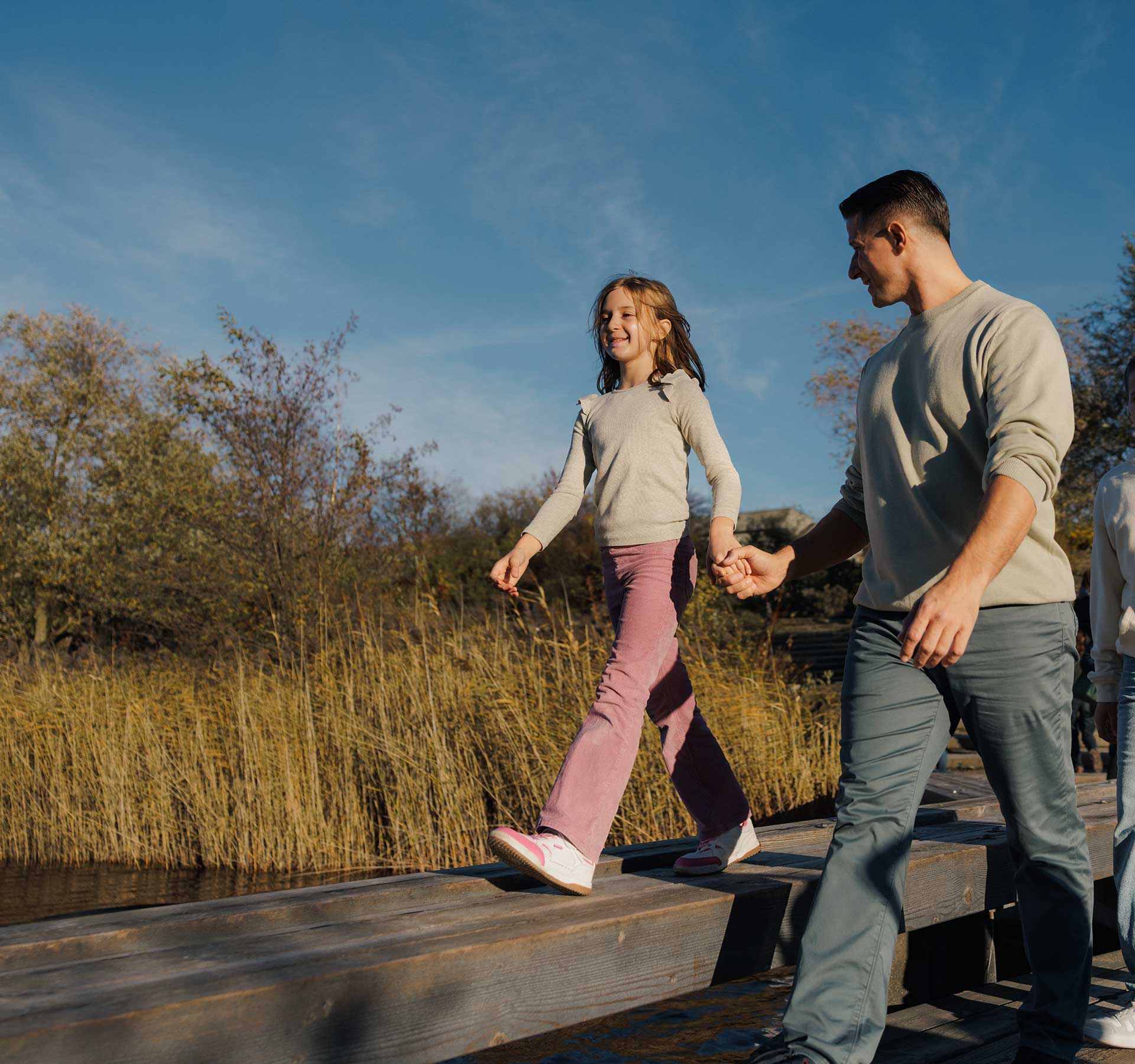 A family is walking across a walkway while the girl balances on a narrower wooden plank.