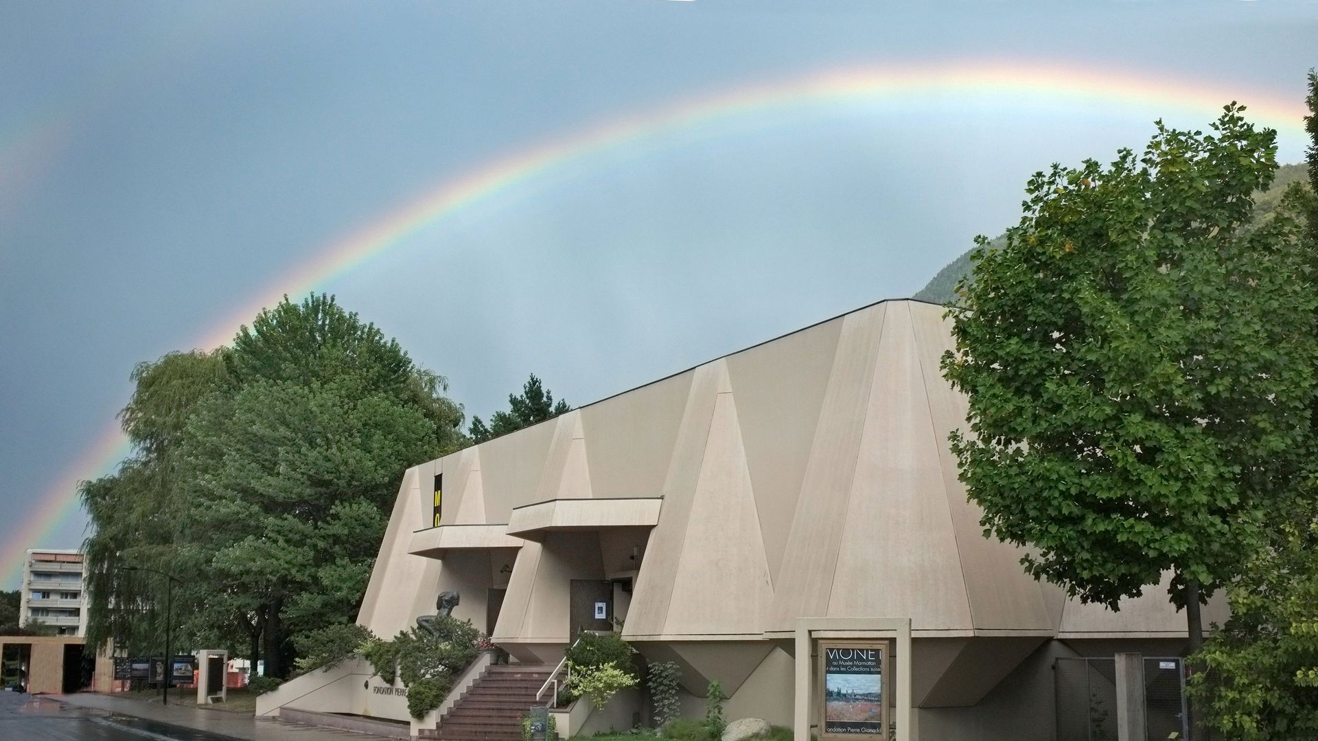 Edificio con scale e alberi, sopra di esso un arcobaleno nel cielo nuvoloso.