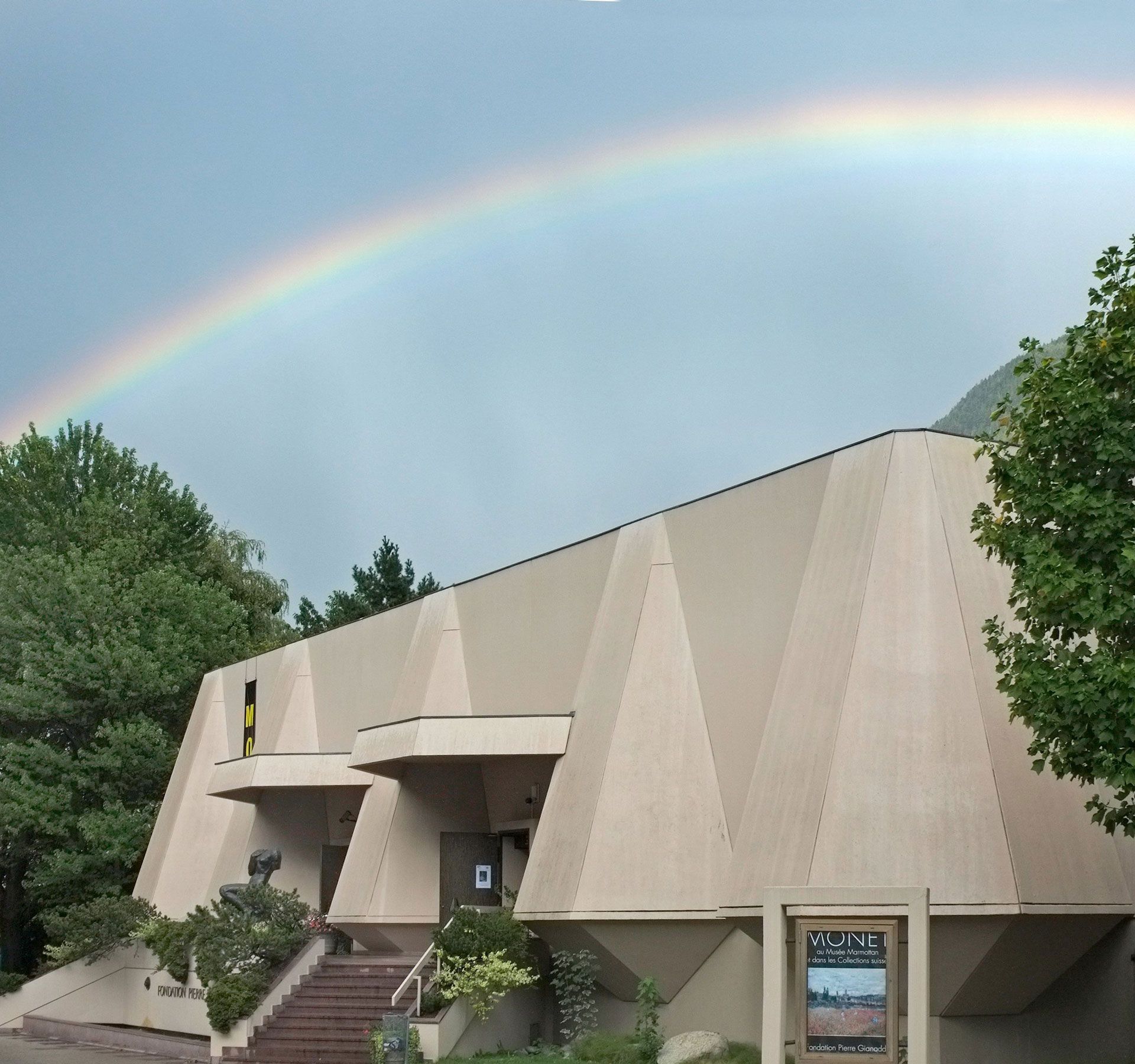 Gebäude mit Treppe und Bäumen, darüber ein Regenbogen am bewölkten Himmel.