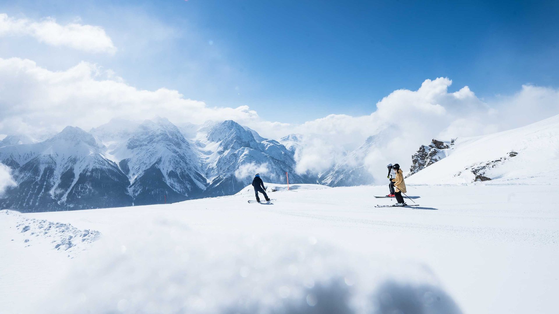 Deux skieurs descendent une pente de montagne enneigée avec des pics montagneux et des nuages en arrière-plan.