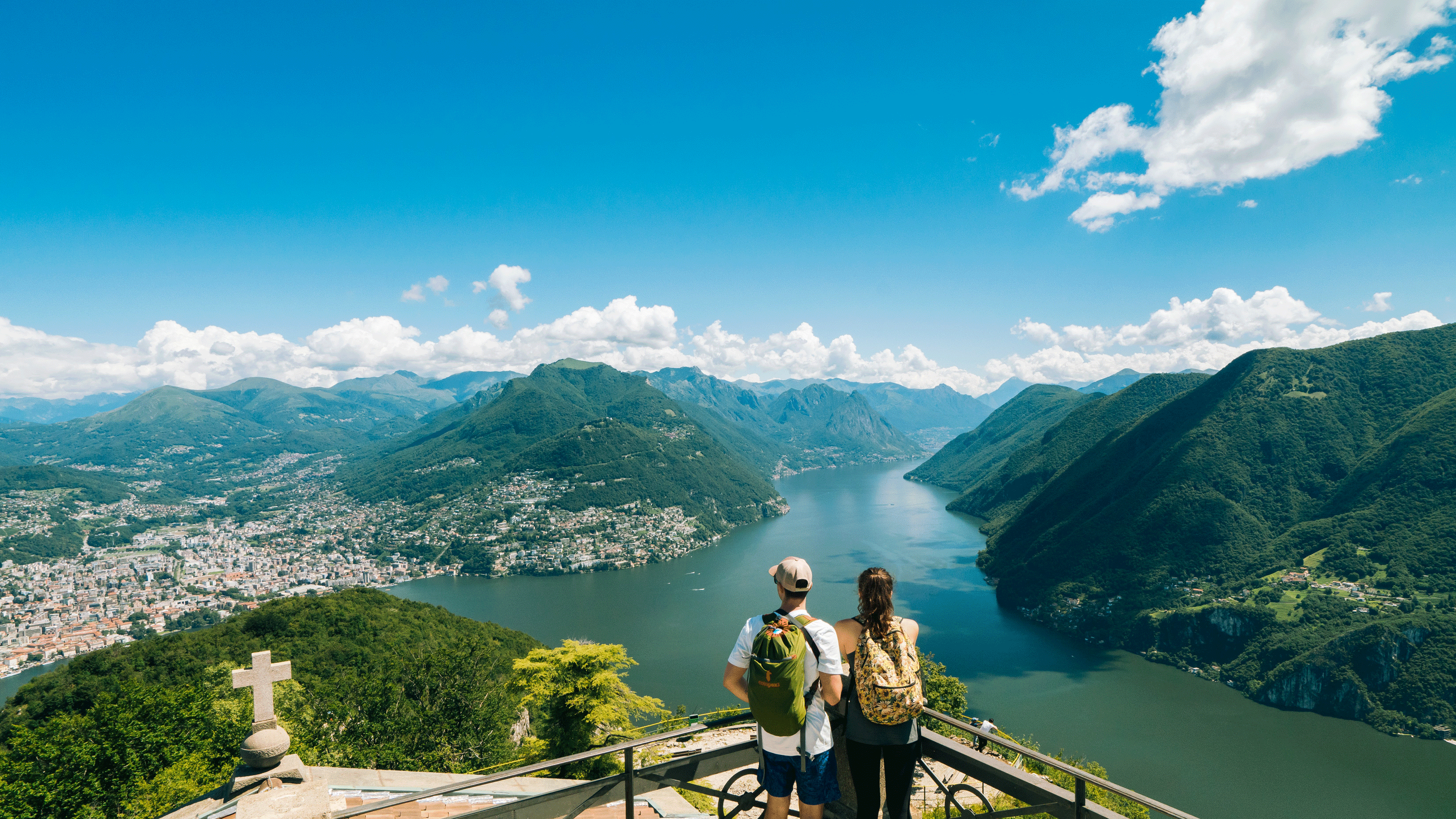 Panoramablick vom Monte San Salvatore auf den von Bergen umgebenen Luganersee mit Besuchern, die die Landschaft von einem hohen Aussichtspunkt aus genießen.