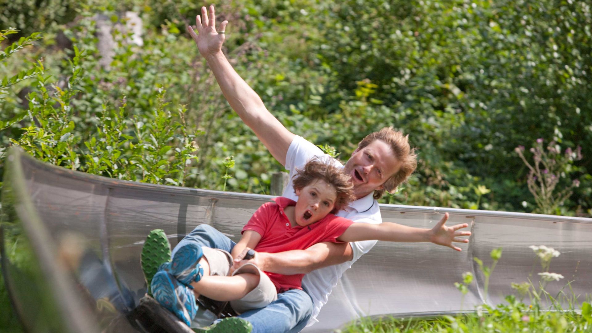 A boy and a man raise an arm in the air as they hurtle down the toboggan run.