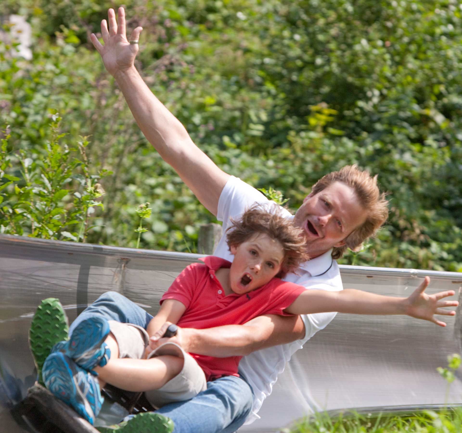 A boy and a man raise an arm in the air as they hurtle down the toboggan run.