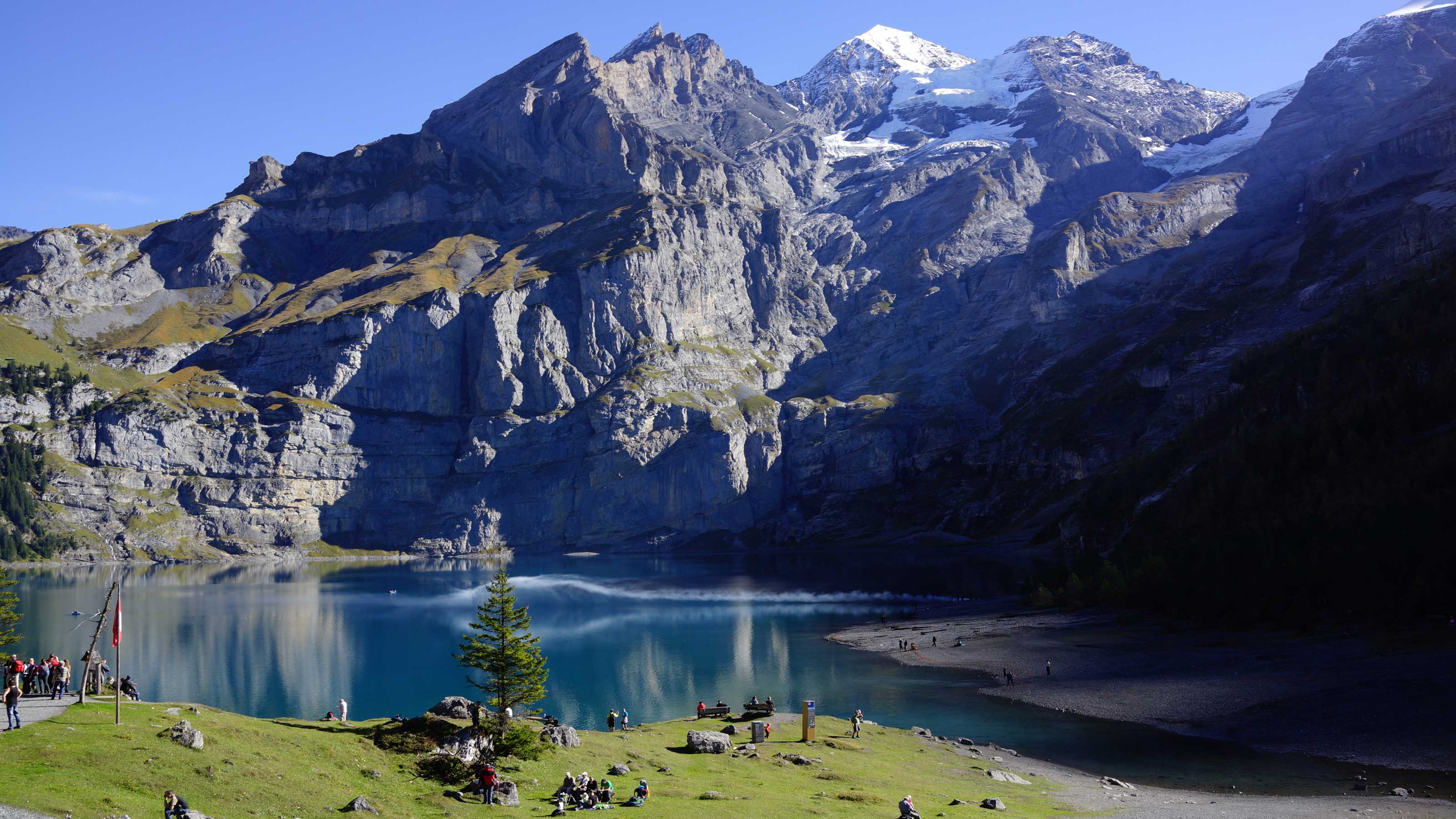 Une vue sur le lac d'Oeschinen. Au premier plan, on voit des gens au bord du lac. En arrière-plan, une montagne enneigée surplombe le lac.