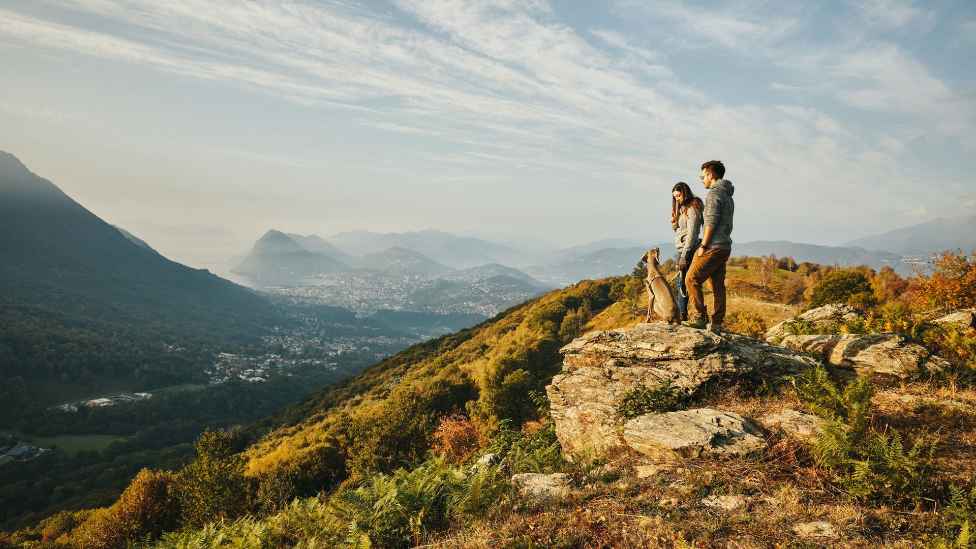 Un uomo e una donna si trovano con il loro cane su una roccia in cima a una piccola montagna. La vista spazia fino a Lugano e al lago. Il sole della sera inonda il paesaggio di una luce brillante e dorata.