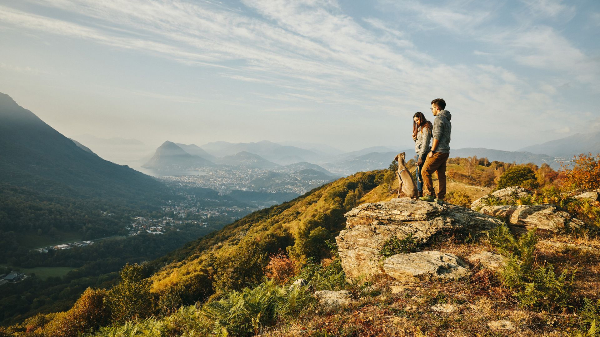 A man and a woman stand with their dog on a rock on a small mountain peak. The view stretches as far as Lugano and the lake. The evening sun bathes the scenery in a bright, golden light.