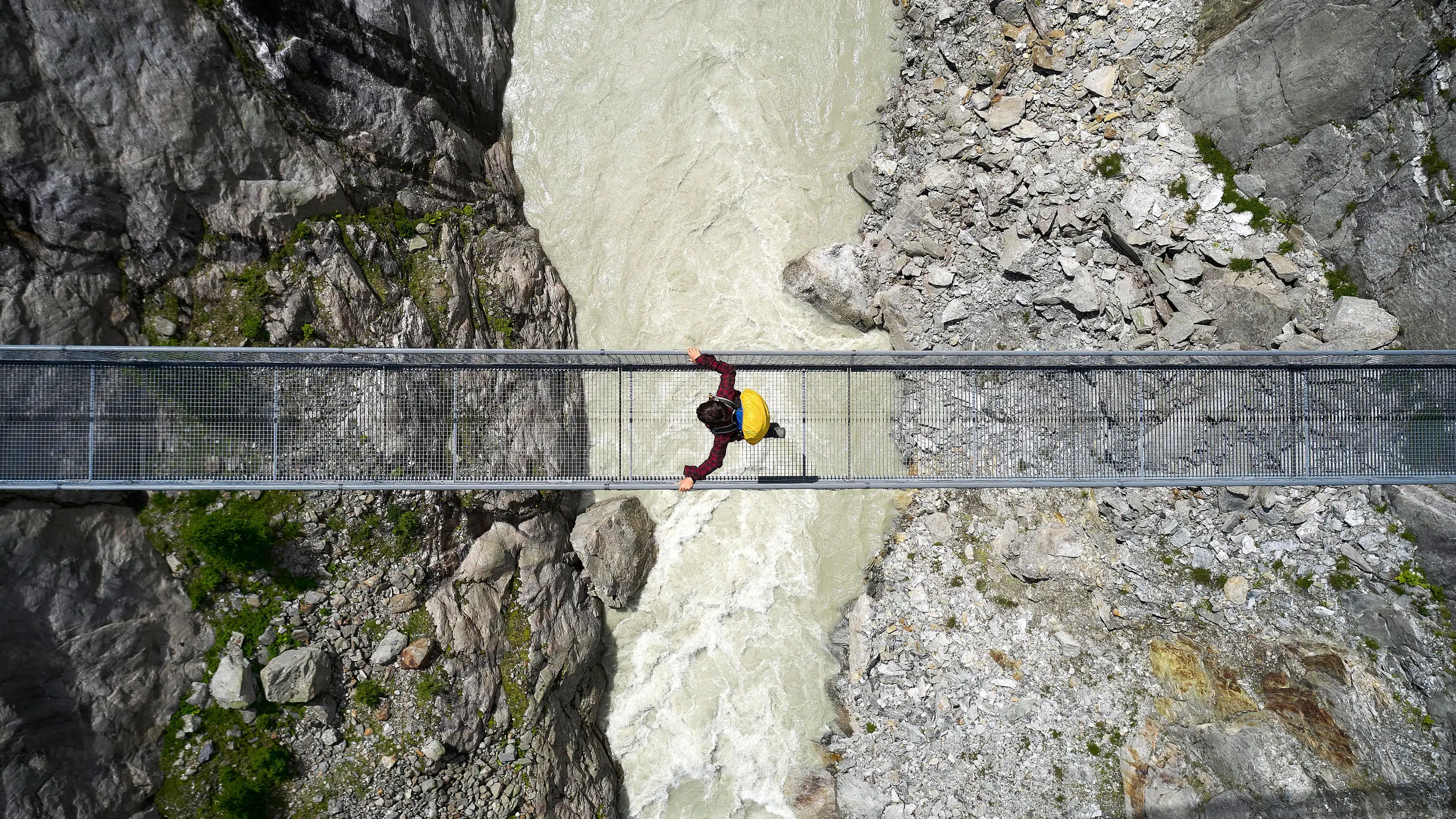  Person mit rotem Hemd und gelbem Rucksack überquert Hängebrücke über reissenden Fluss in felsiger Schlucht