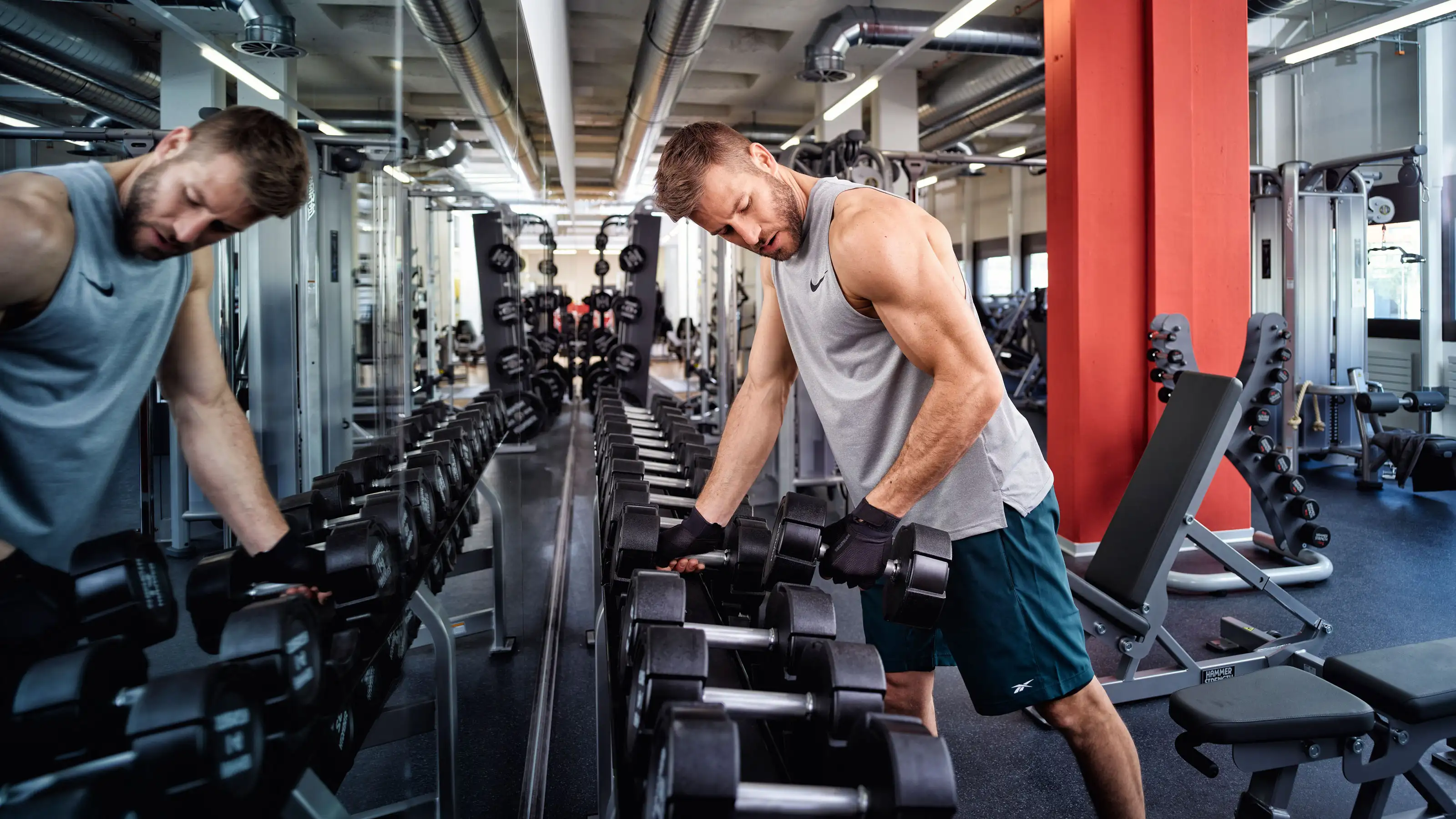 Mann in Sportkleidung hebt Kurzhantel vor Spiegel in Fitnessstudio.