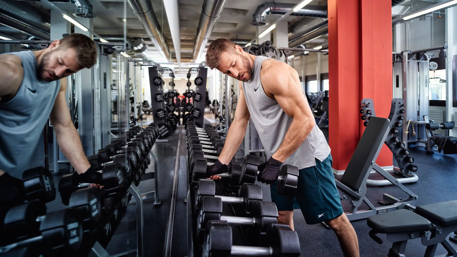 Mann in Sportkleidung hebt Kurzhantel vor Spiegel in Fitnessstudio.
