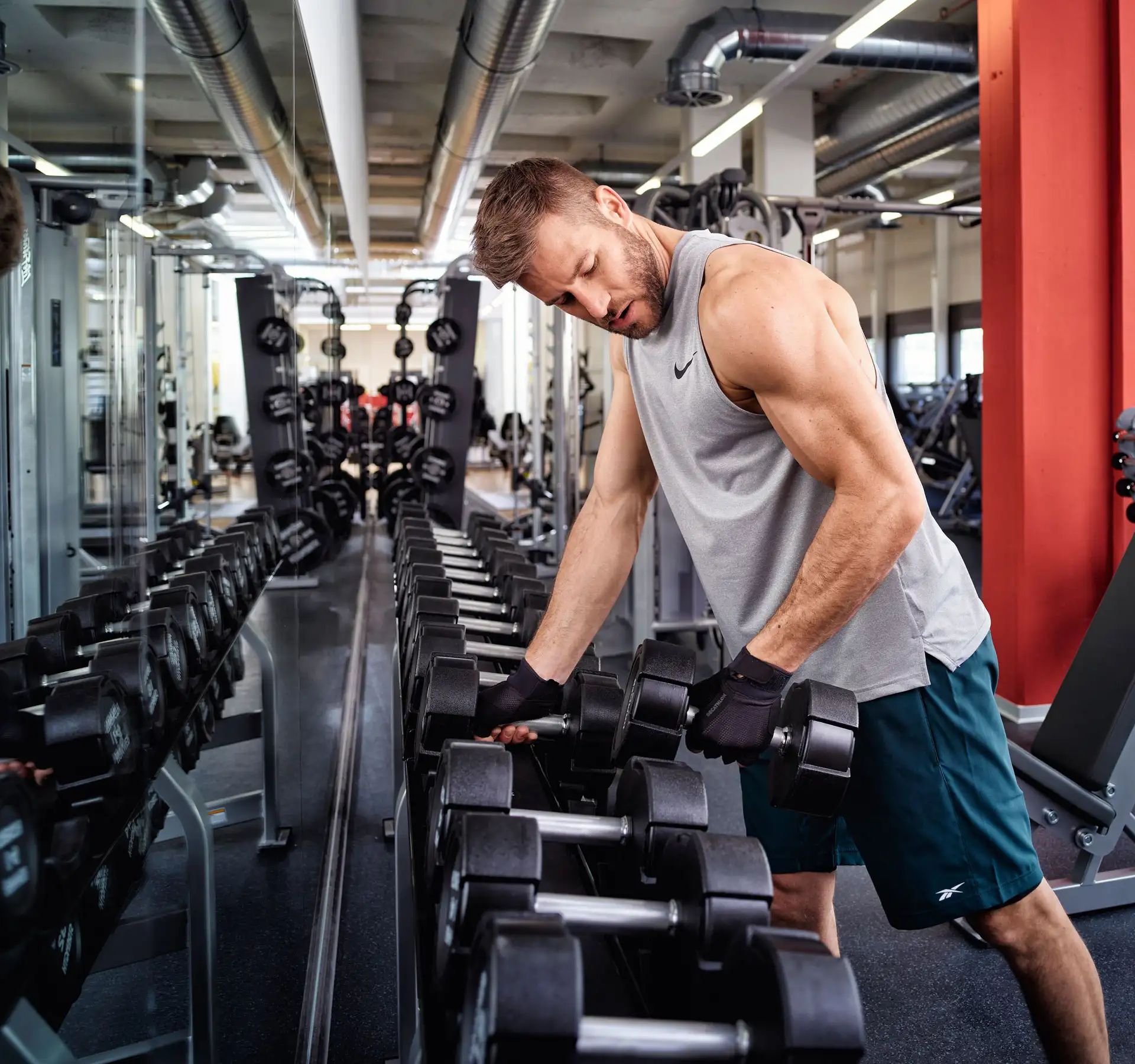Mann in Sportkleidung hebt Kurzhantel vor Spiegel in Fitnessstudio.