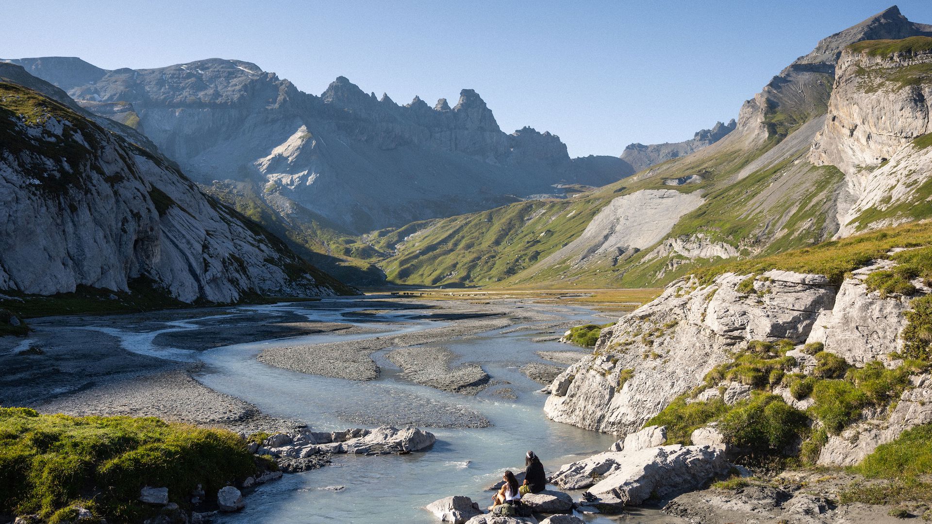 Two people sit on a stone, enjoying the expansive view of the mountain panorama and the river.