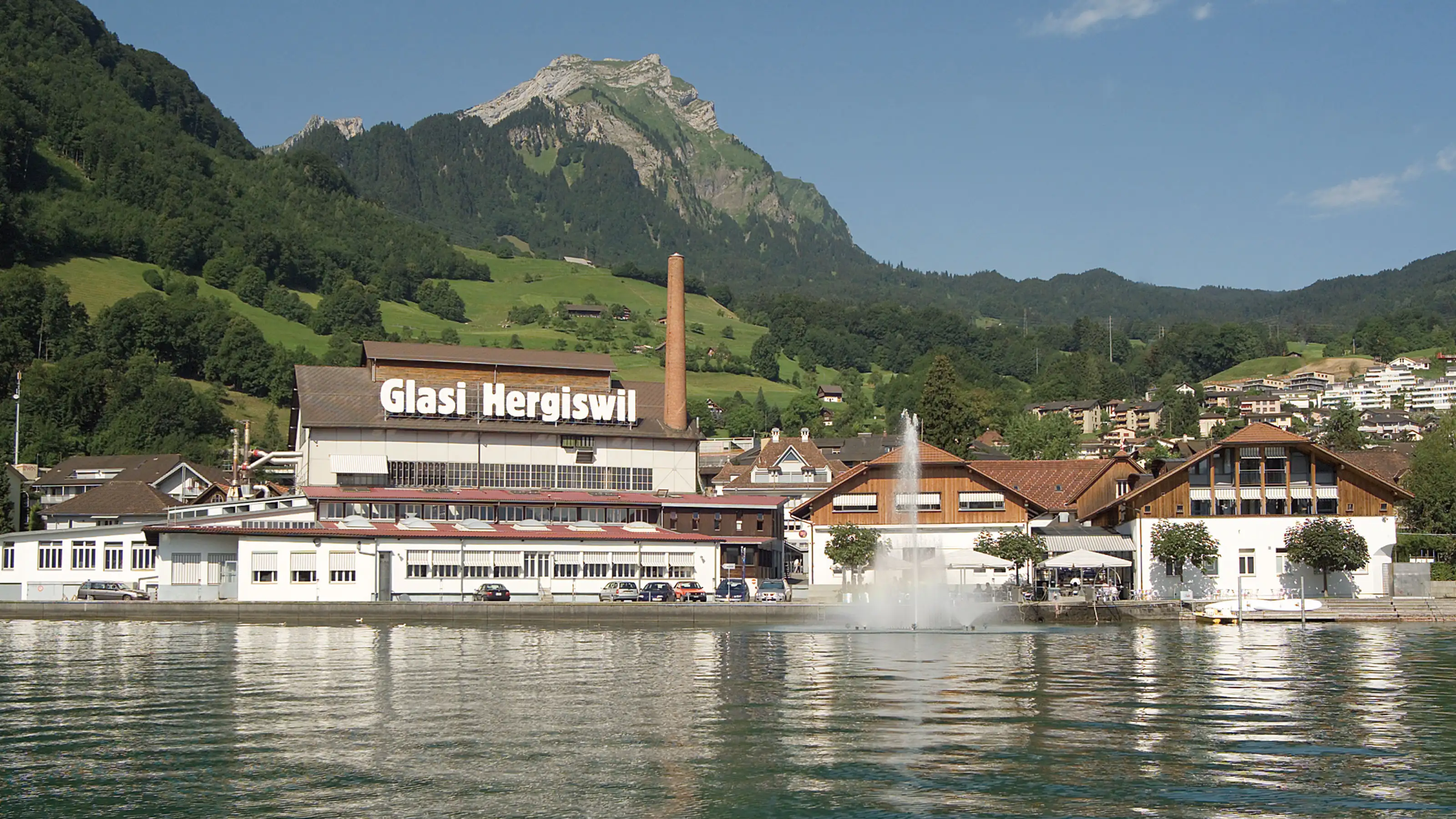 La Glasi Hergiswil est située directement au bord du lac, entourée de collines verdoyantes et de montagnes, avec un bâtiment industriel emblématique, une cheminée et une fontaine au premier plan.