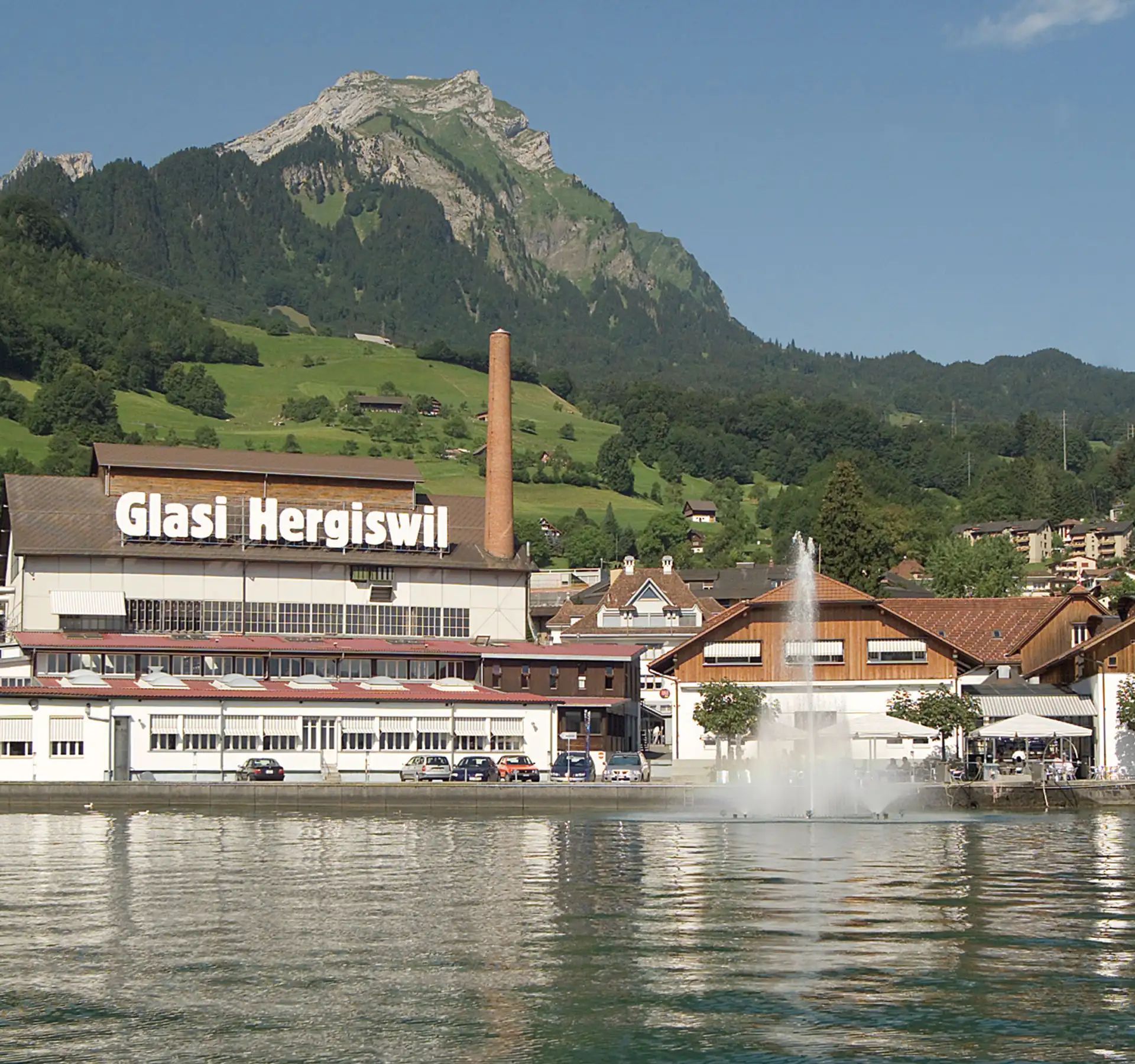 La Glasi Hergiswil est située directement au bord du lac, entourée de collines verdoyantes et de montagnes, avec un bâtiment industriel emblématique, une cheminée et une fontaine au premier plan.