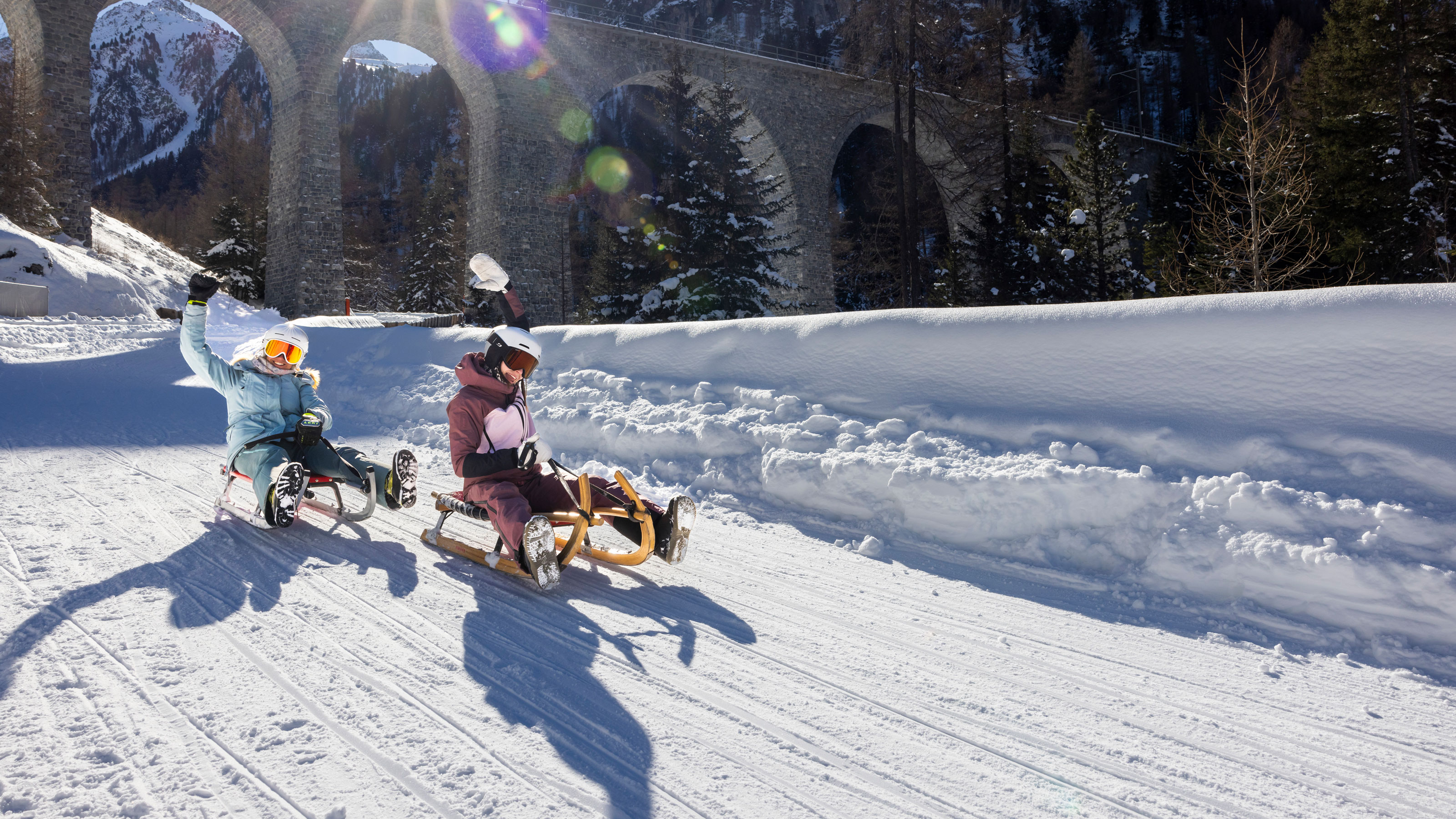 Deux personnes faisant de la luge sur un chemin enneigé avec un pont en arrière-plan.