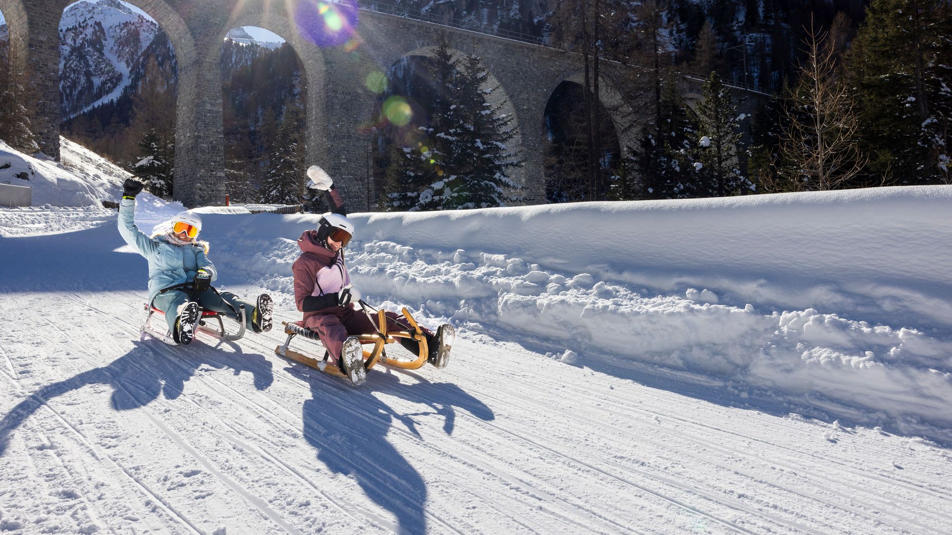 Two people sledding on a snowy path with a bridge in the background.