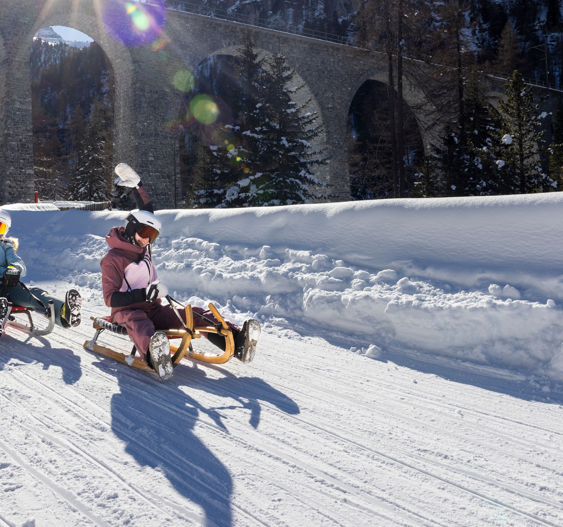 Two people sledding on a snowy path with a bridge in the background.