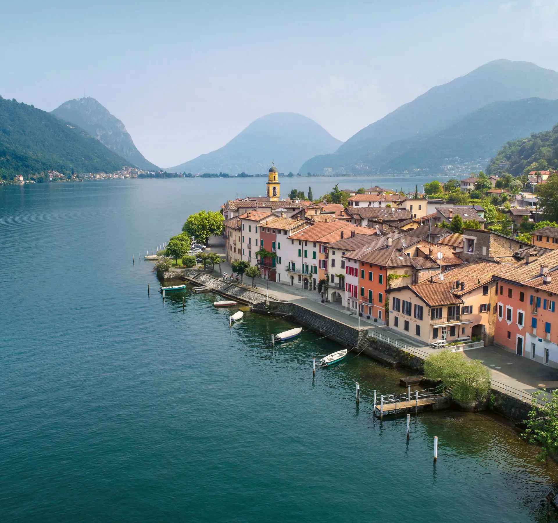 Uferpromenade mit bunten Häusern und kleinen Booten am Seeufer im Tessin umgeben von bewaldeten Bergen.