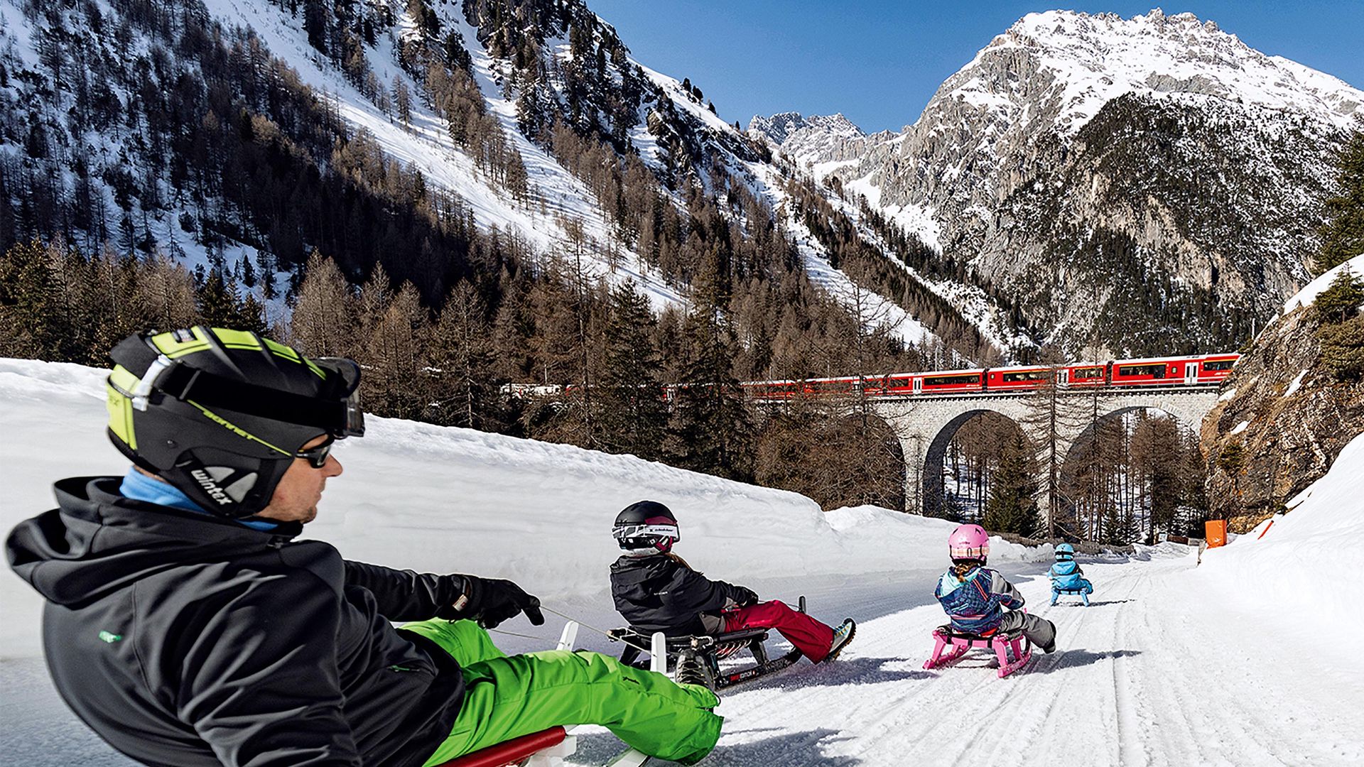 A man and 3 children are sledging down the fast sledge run towards a railway bridge where a Rhaetian Railway train is just crossing it.