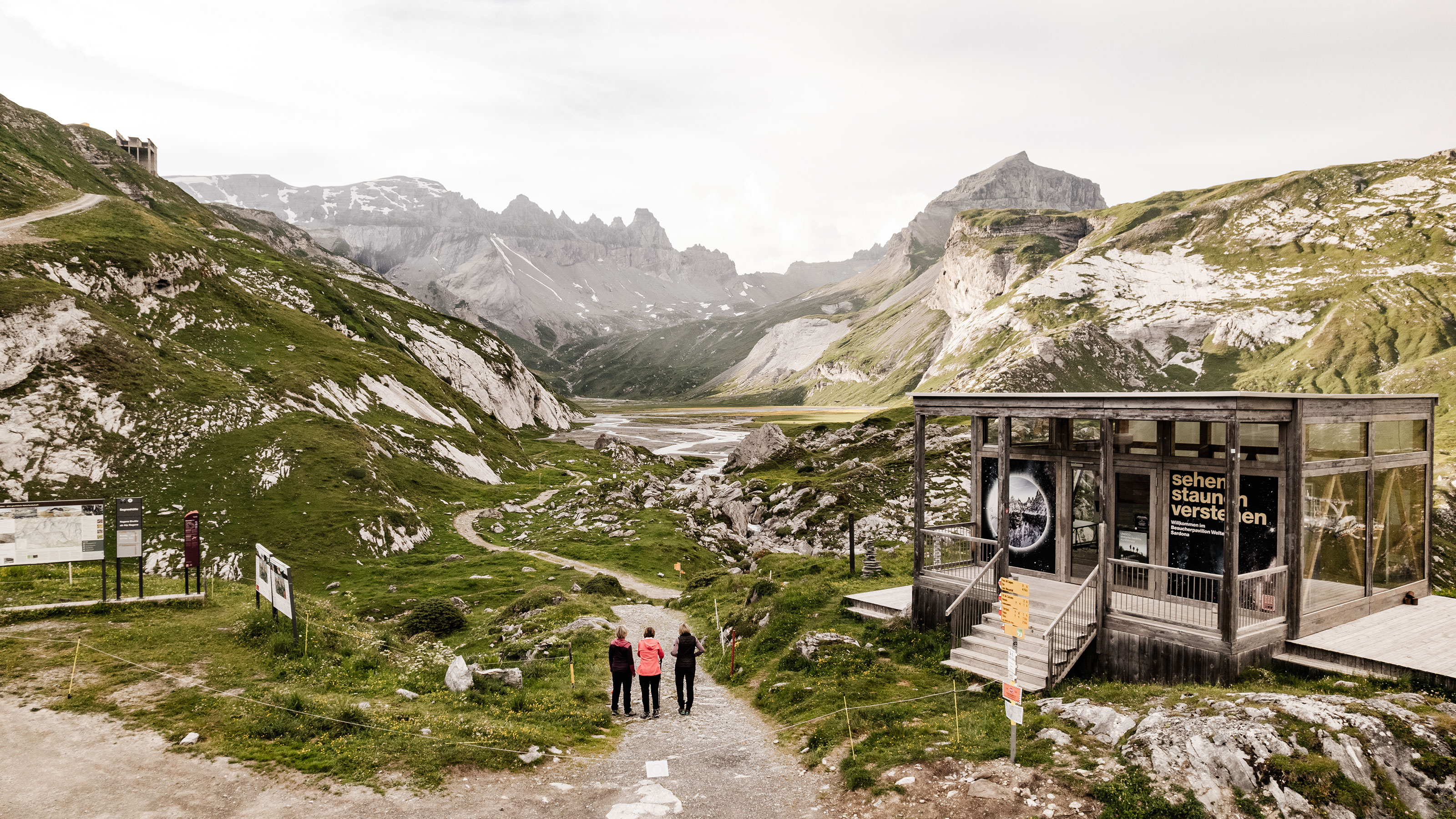 Blick auf das UNESCO-Welterbe Tektonikarena Sardona in der Schweiz: Eine Gruppe von drei Wandernden steht auf einem Weg, der durch eine beeindruckende alpine Landschaft führt.