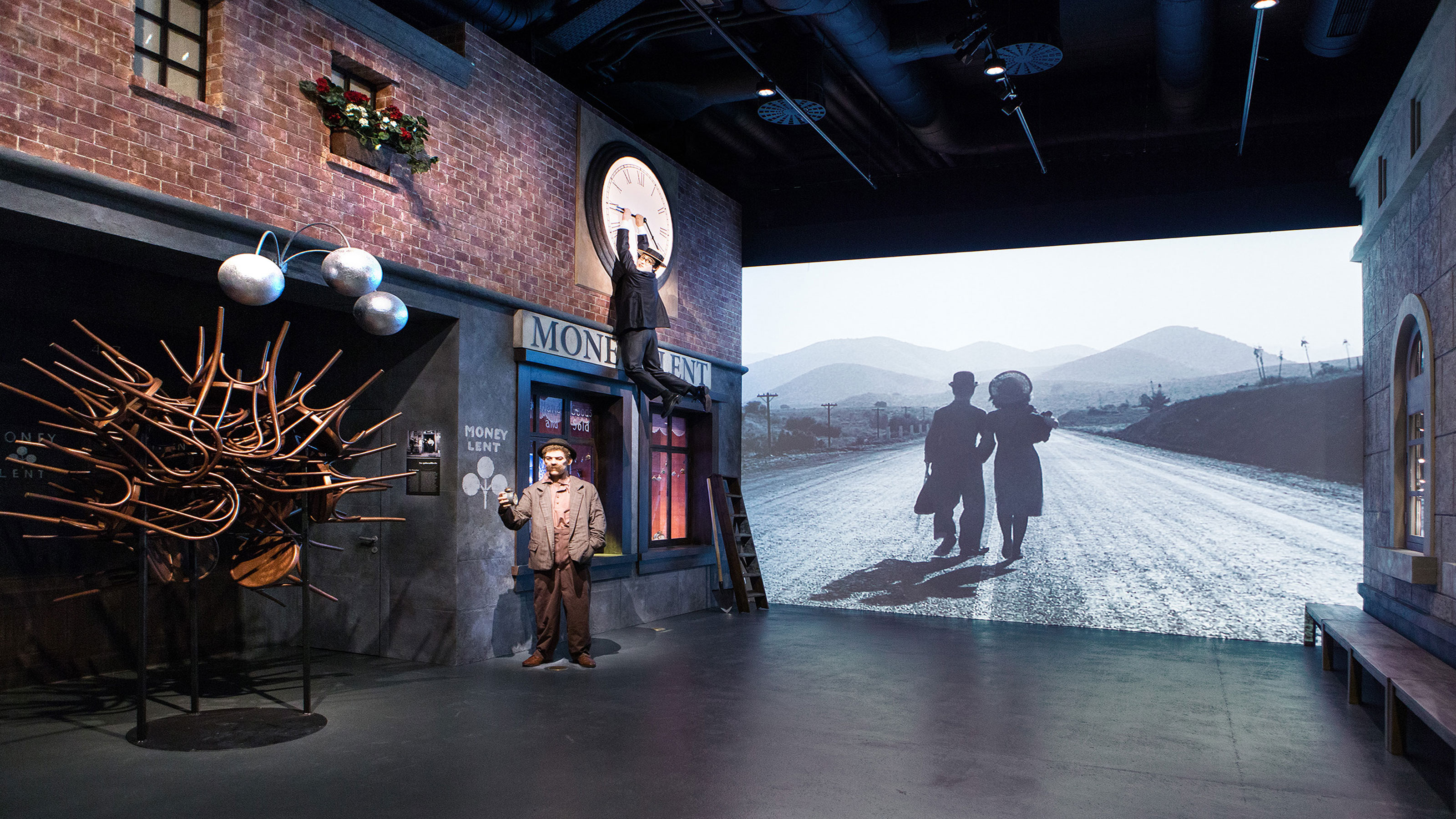 Dans une exposition de musée, on voit un bâtiment en briques, une poupée déguisée en Charlie Chaplin dans une rue et un film en noir et blanc dans lequel deux personnes marchent vers un horizon montagneux.