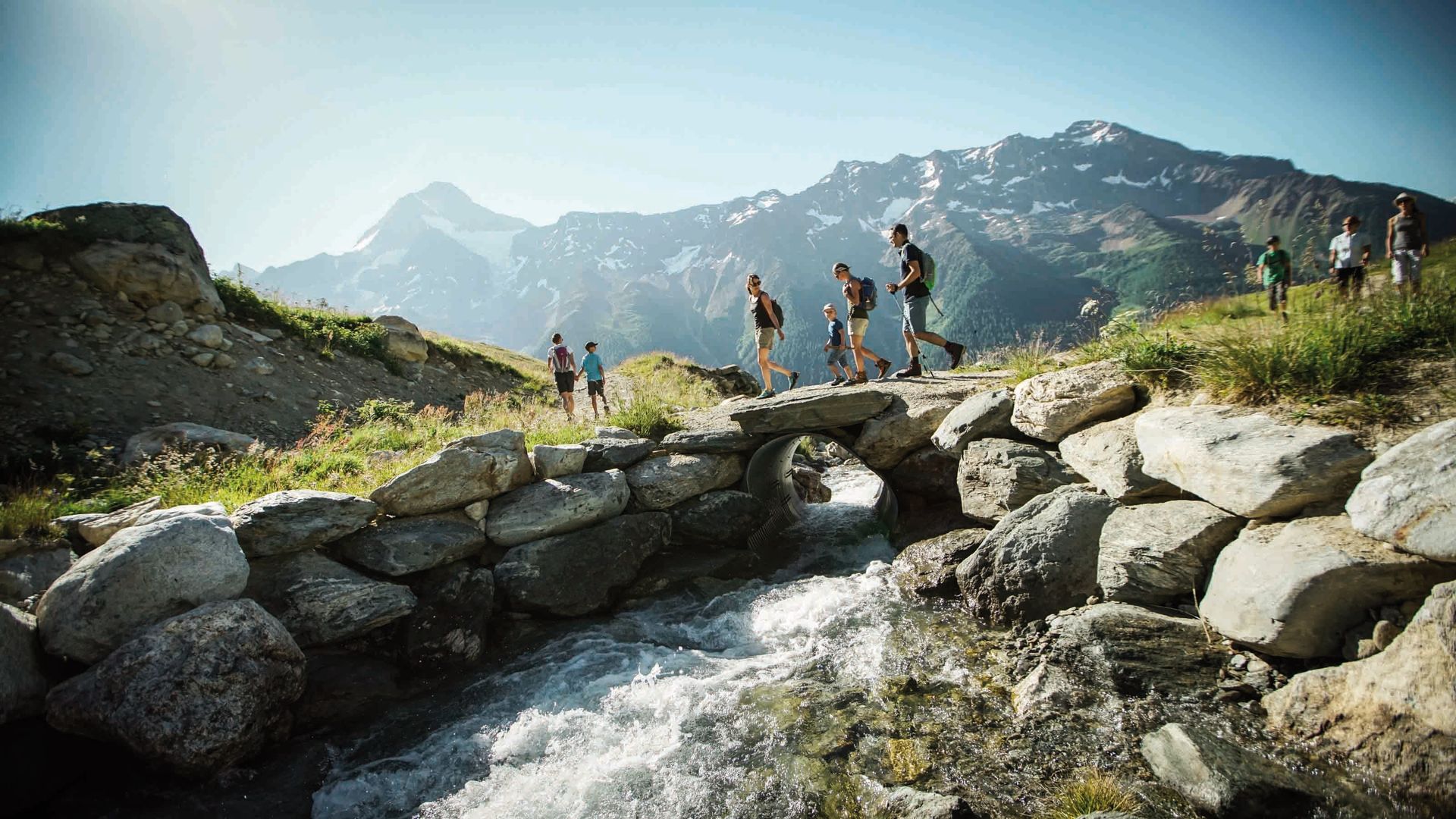 Eine Familie wandert über eine kleine Steinbrücke über einem Bach auf dem Lötschentaler Höhenweg.
