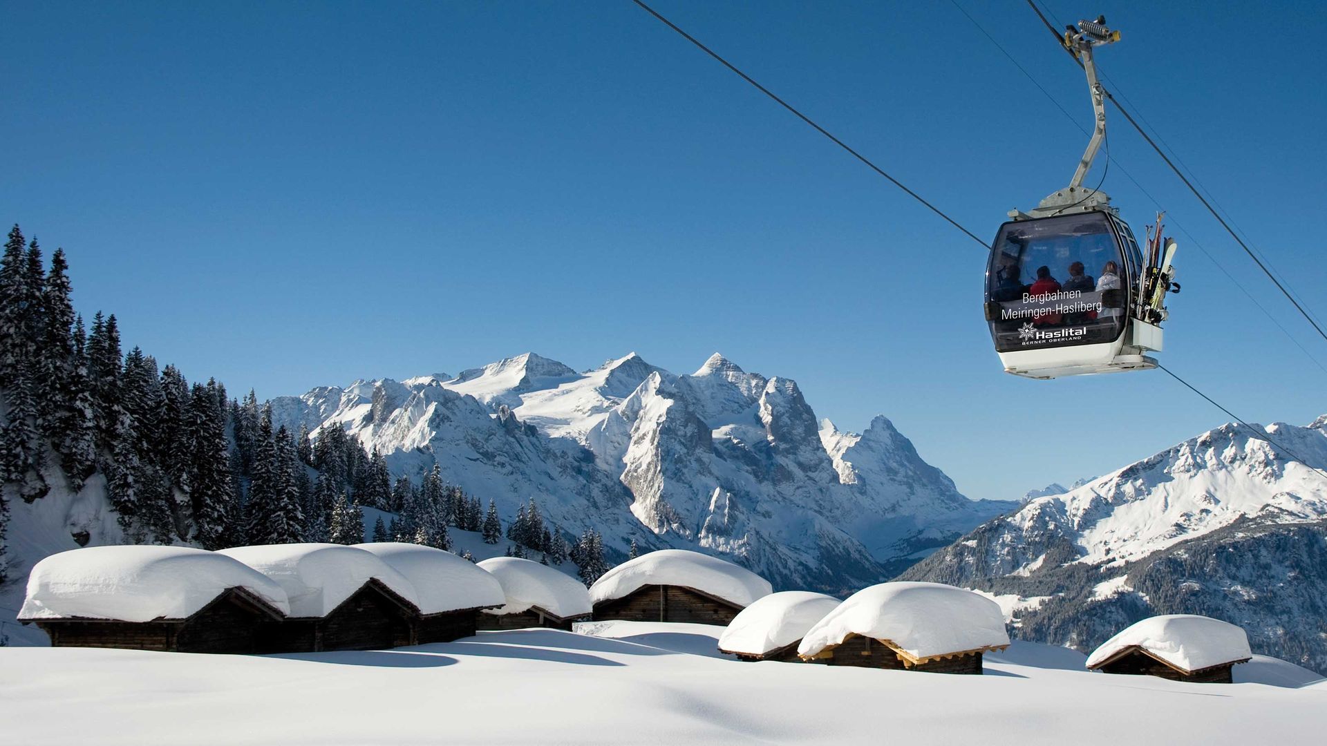 Gondola lift in the Meiringen-Hasliberg ski resort floats above snow-covered huts with pine forests and mountain panoramas in the background.