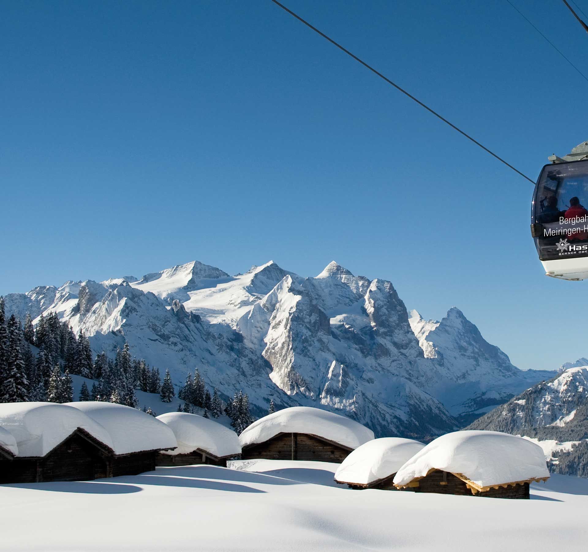 La télécabine du domaine skiable de Meiringen-Hasliberg survole des chalets enneigés, avec une forêt de sapins et un panorama montagneux en arrière-plan.