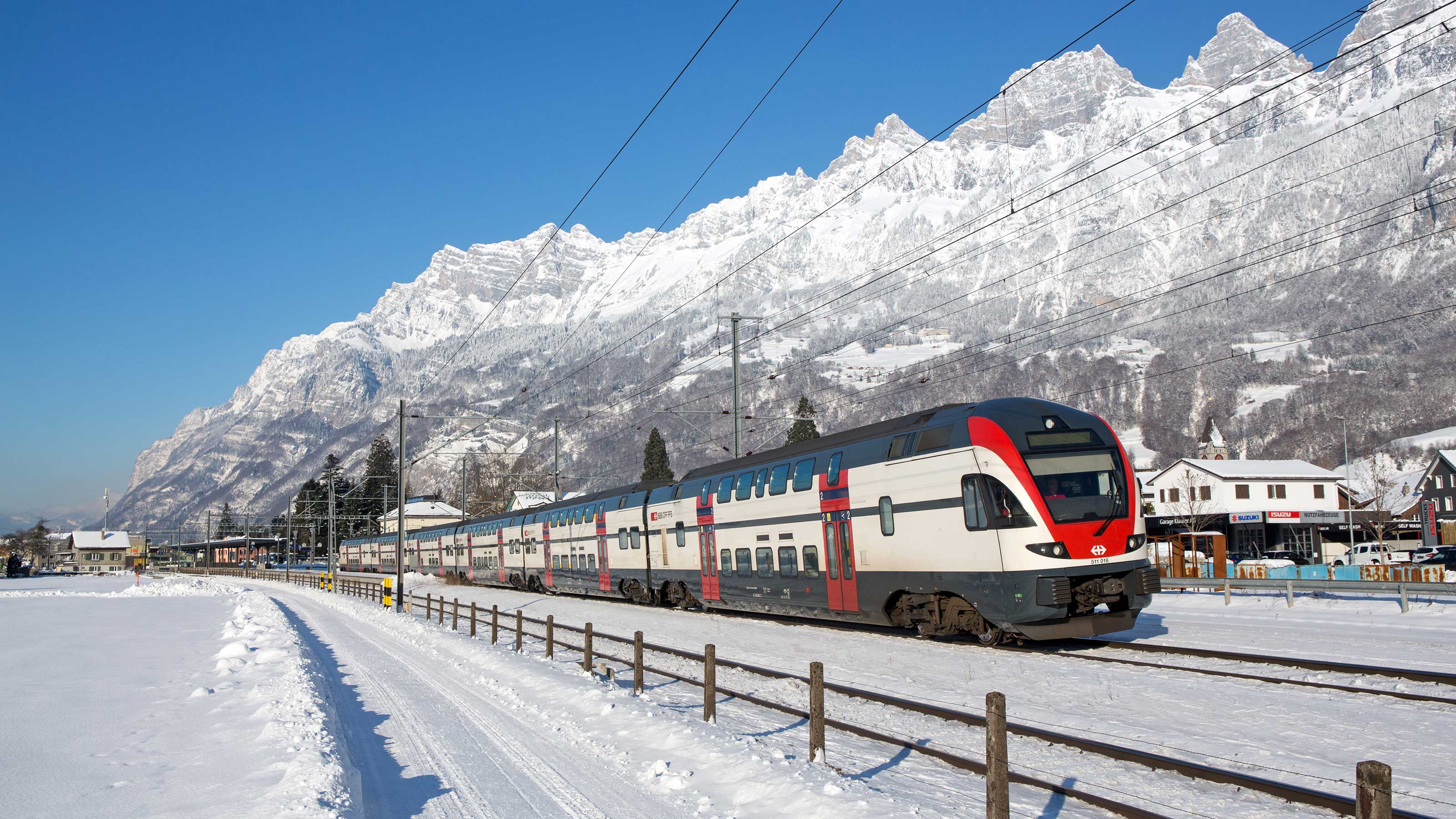 An SBB train during its journey through a snow-covered village. A white mountain range stretches out in the background.