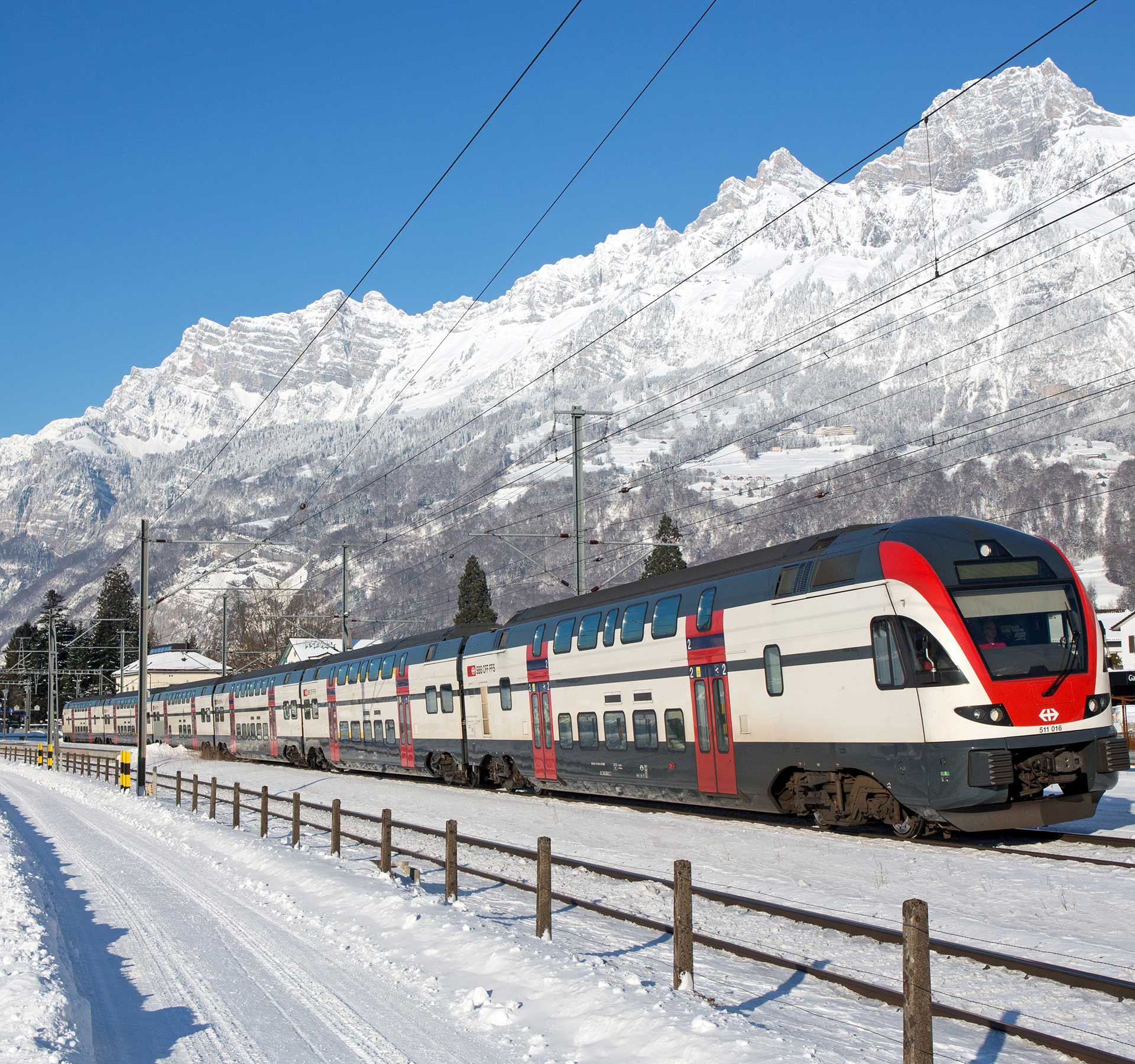 Ein SBB Zug während seiner Fahr durch ein Verschneeeites Dorf. Im Hintergrund erstreckt sich eine ebenfalls weisse Bergkette.