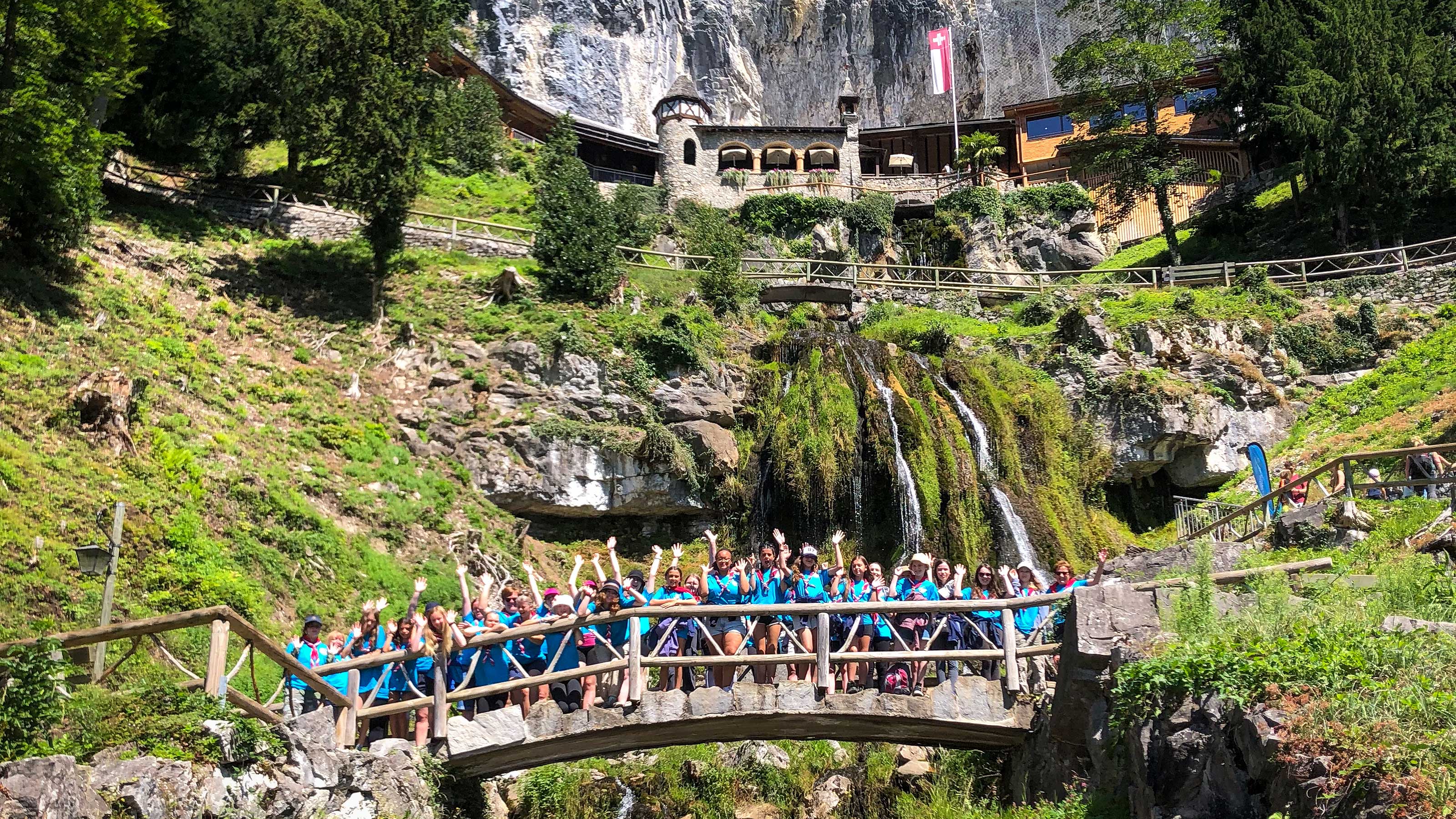 Exterior view of the Beatus Caves. On an imposing rock face, a tower made of stone stands next to a new building made of wood. Below, a small waterfall flows down the rock face.