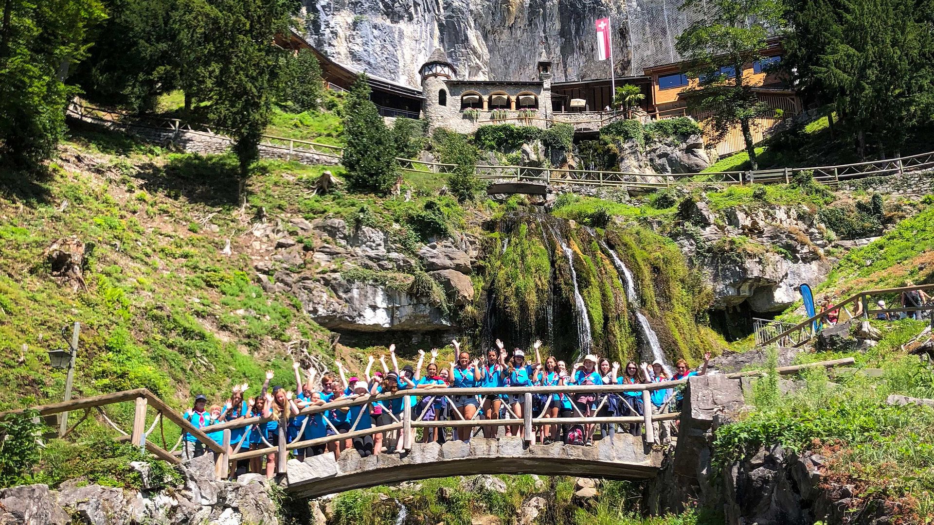 Exterior view of the Beatus Caves. On an imposing rock face, a tower made of stone stands next to a new building made of wood. Below, a small waterfall flows down the rock face.