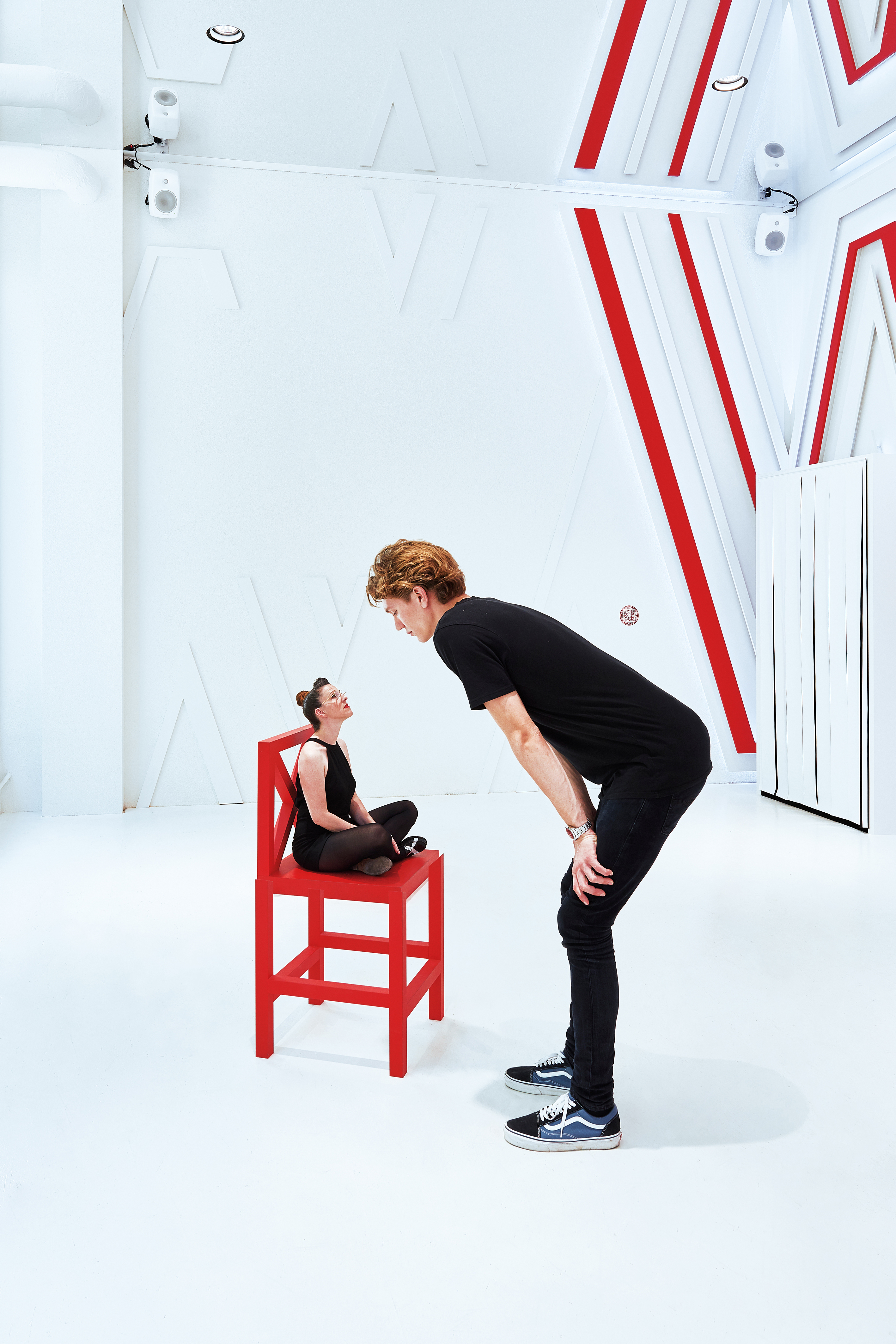 A woman sits cross-legged on a red chair. In front of it stands a guy who, with his apparent oversize, looks down on the woman in the chair from above. This is an optical illusion.
