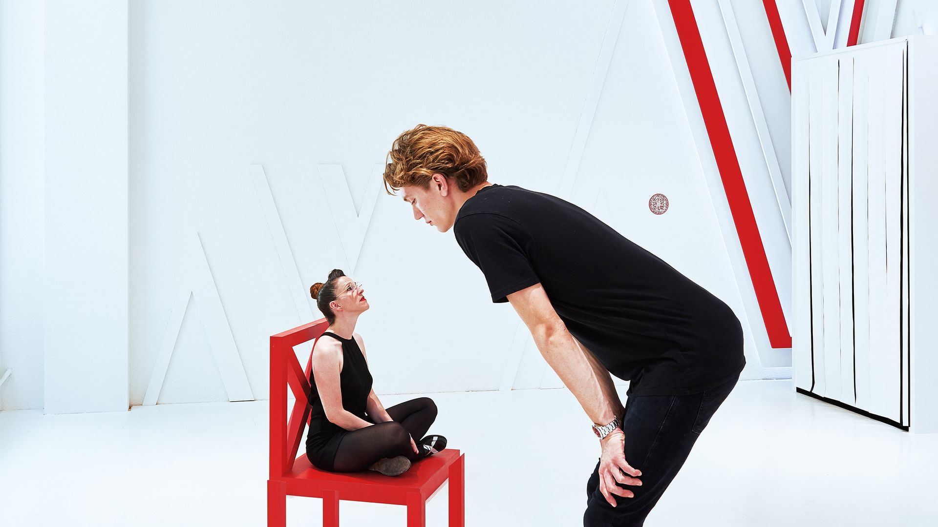 Eine Frau sitzt im Schneidersitz auf einem roten Stuhl. Davor steht ein Typ welcher mit seiner scheinbaren Übergrösse von oben auf die Frau auf dem Stuhl hinab blickt. Es handelt sich um eine optische Täuschung.