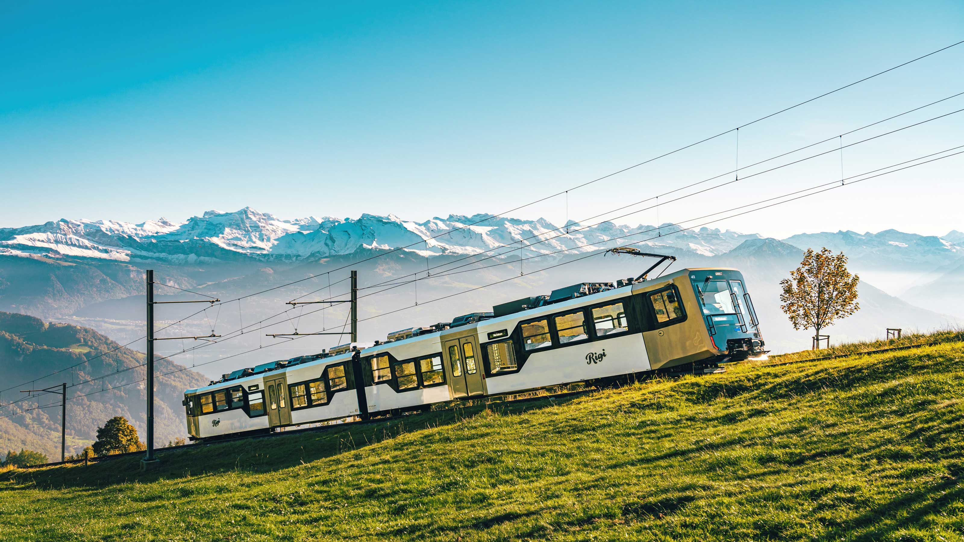 Un train blanc du Rigi en route vers le sommet. En arrière-plan, on aperçoit un magnifique panorama de montagnes.