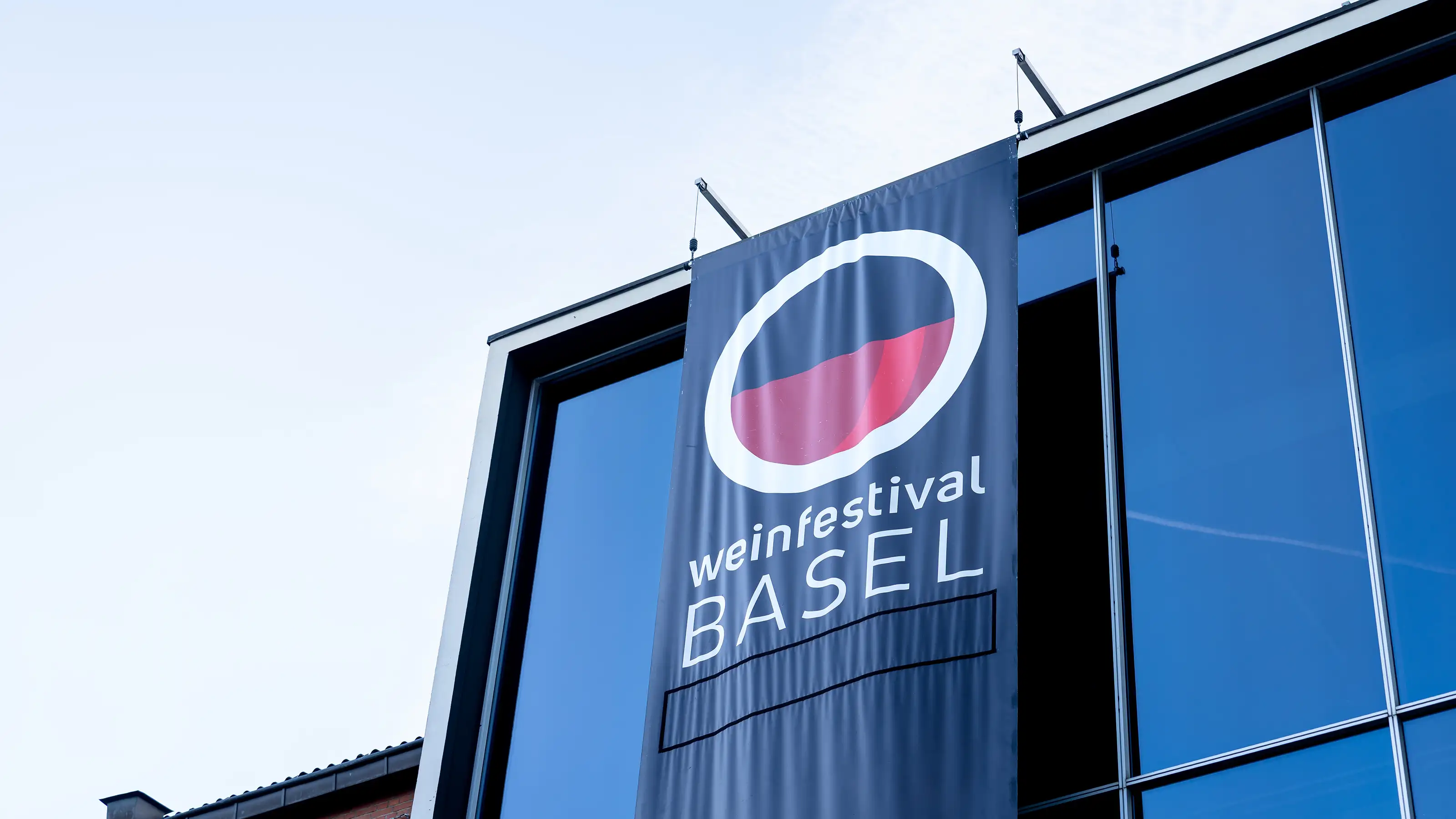 Fahne mit rundem Logo und Schriftzug weinfestival Basel an einem modernen Glasgebäude.