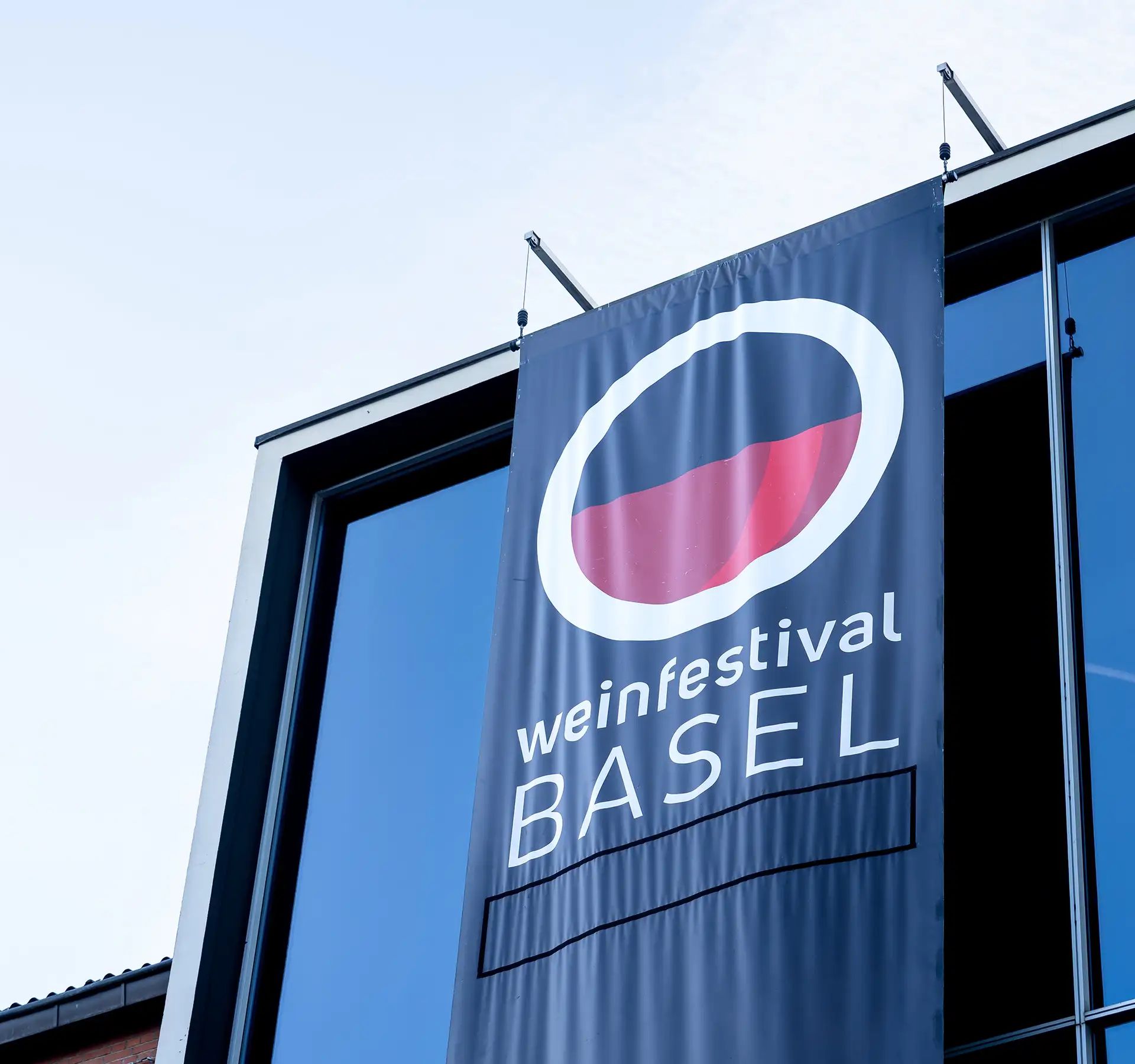 Fahne mit rundem Logo und Schriftzug weinfestival Basel an einem modernen Glasgebäude.