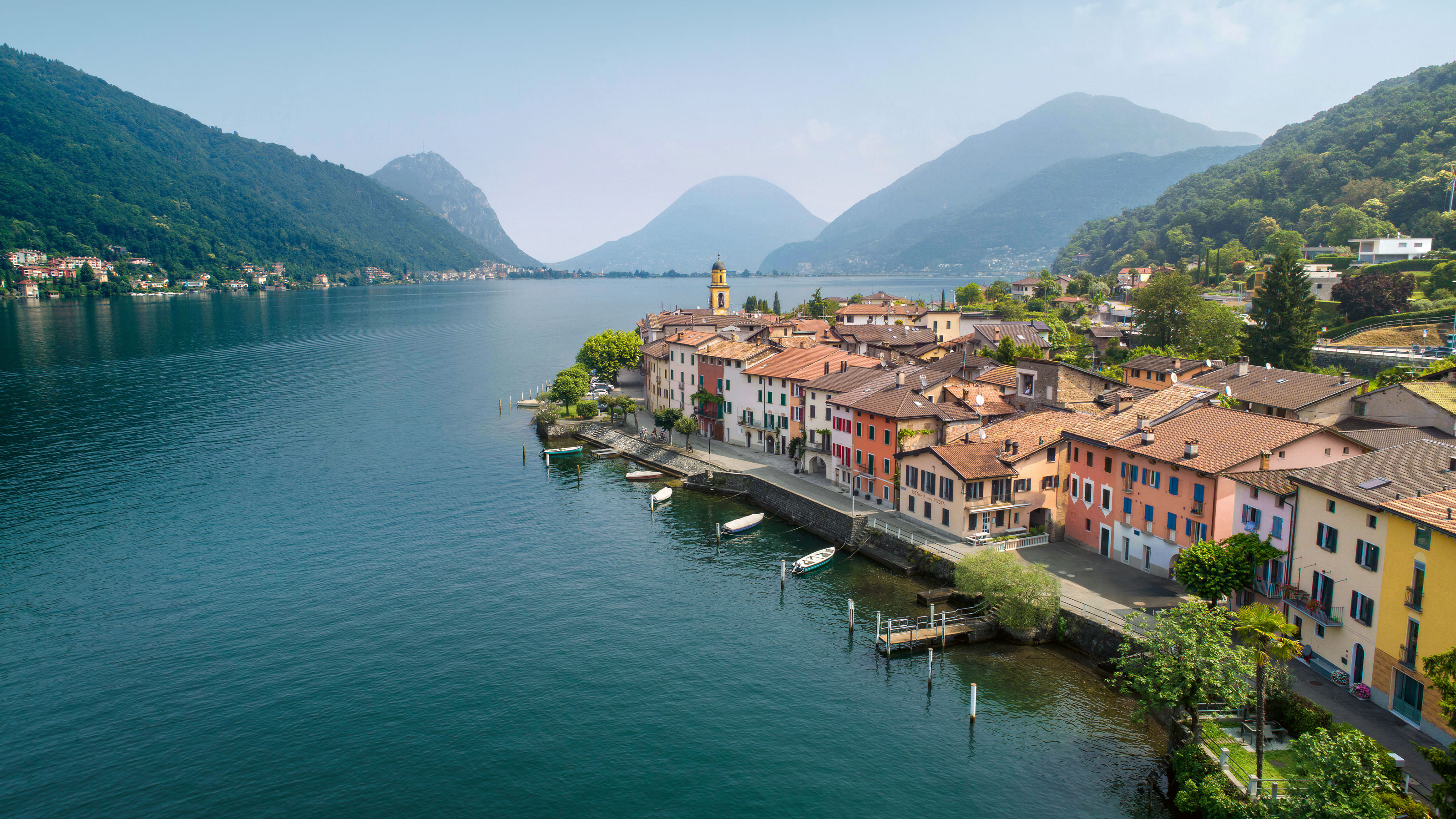 Waterfront promenade with colorful houses and small boats along the lakeshore in Ticino surrounded by forested mountains.