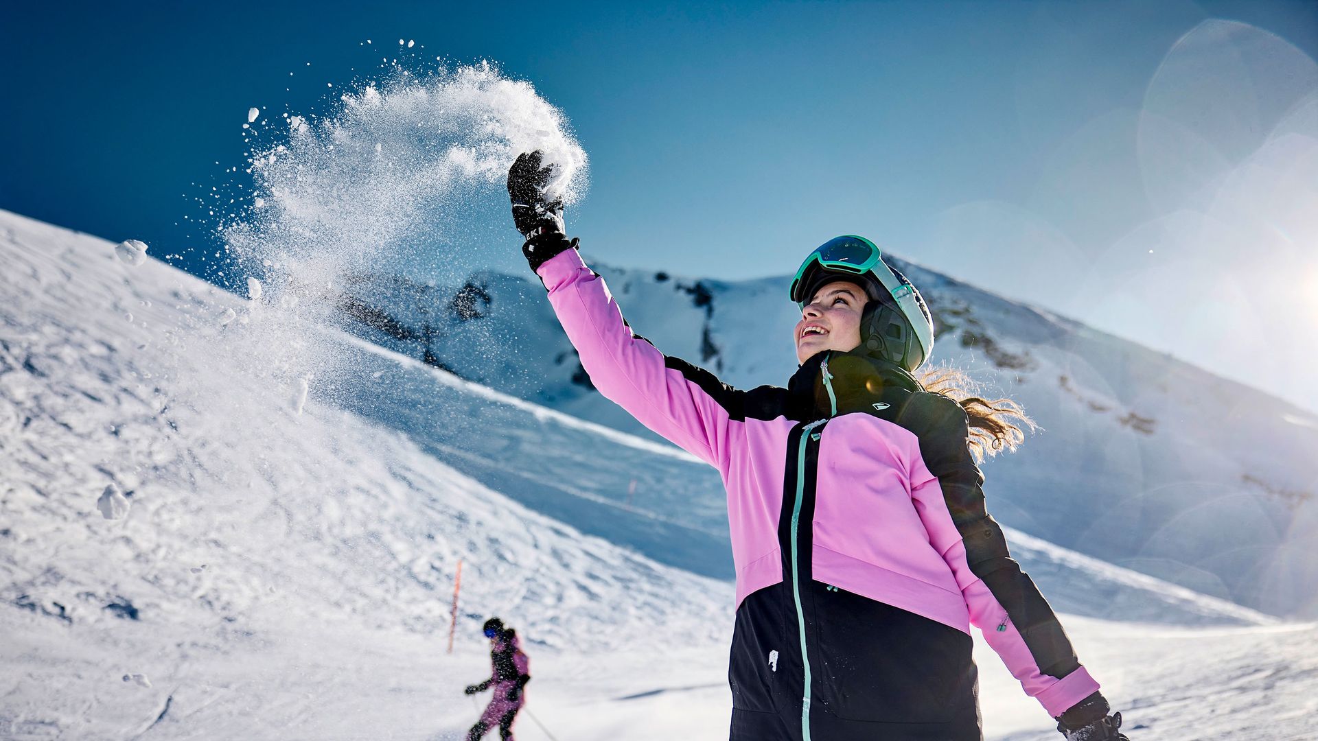 Mädchen in rosa-schwarzer Skijacke wirft Schnee in die Luft auf der Klewenalp-Stockhütte, Berg im Hintergrund.