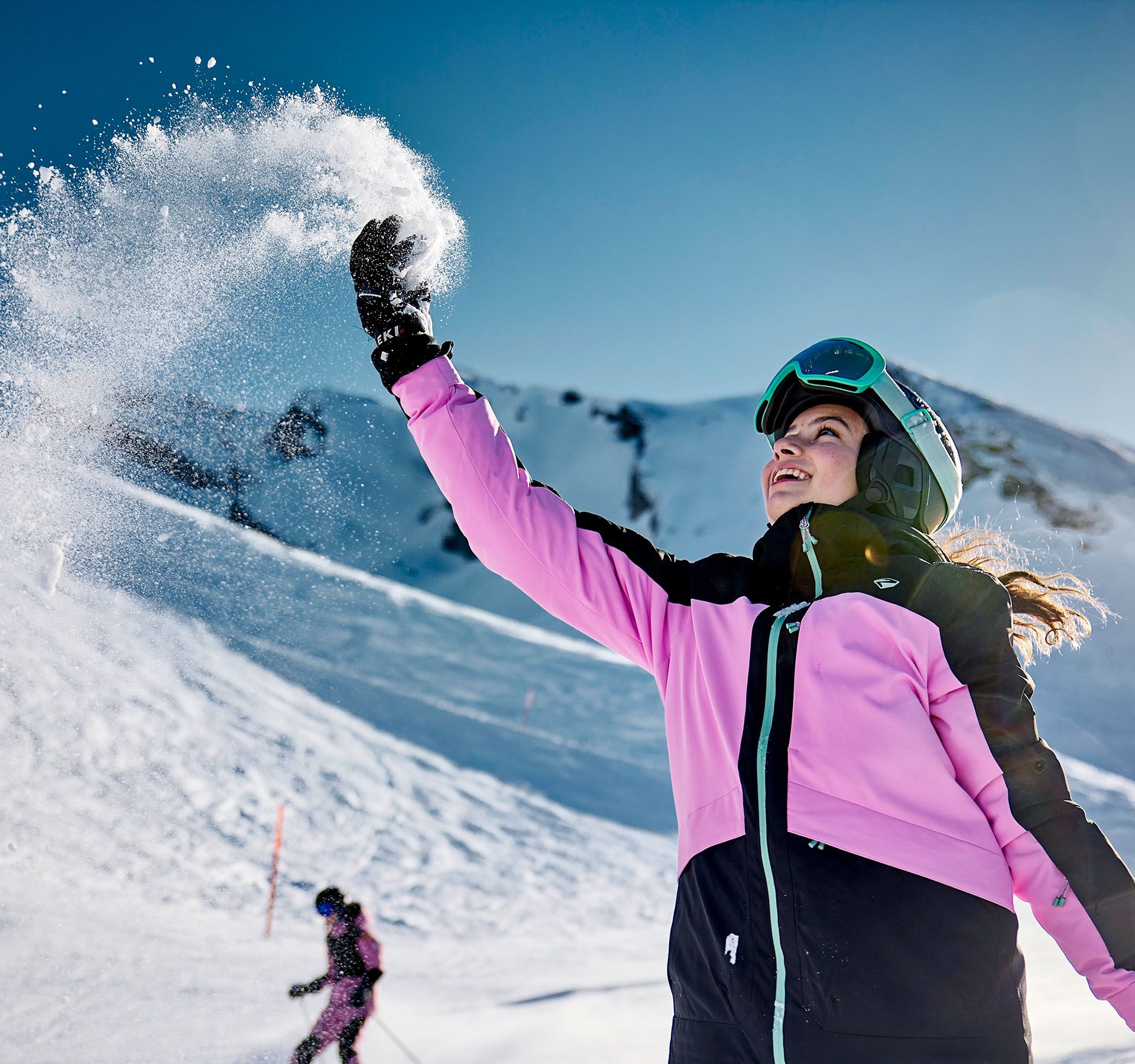 Girl in pink and black ski jacket throwing snow into the air at Klewenalp-Stockhütte ski area, mountain in background.