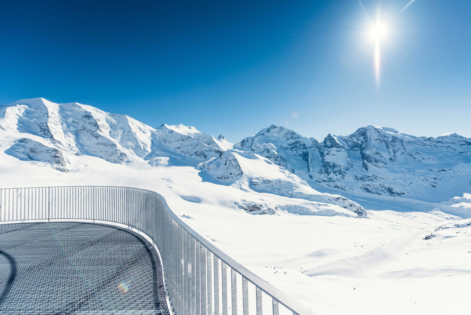 Ein Panoramablick auf schneebedeckte Berge bei strahlendem Sonnenschein mit einem gebogenen Metallzaun im Vordergrund an einem klaren Tag.