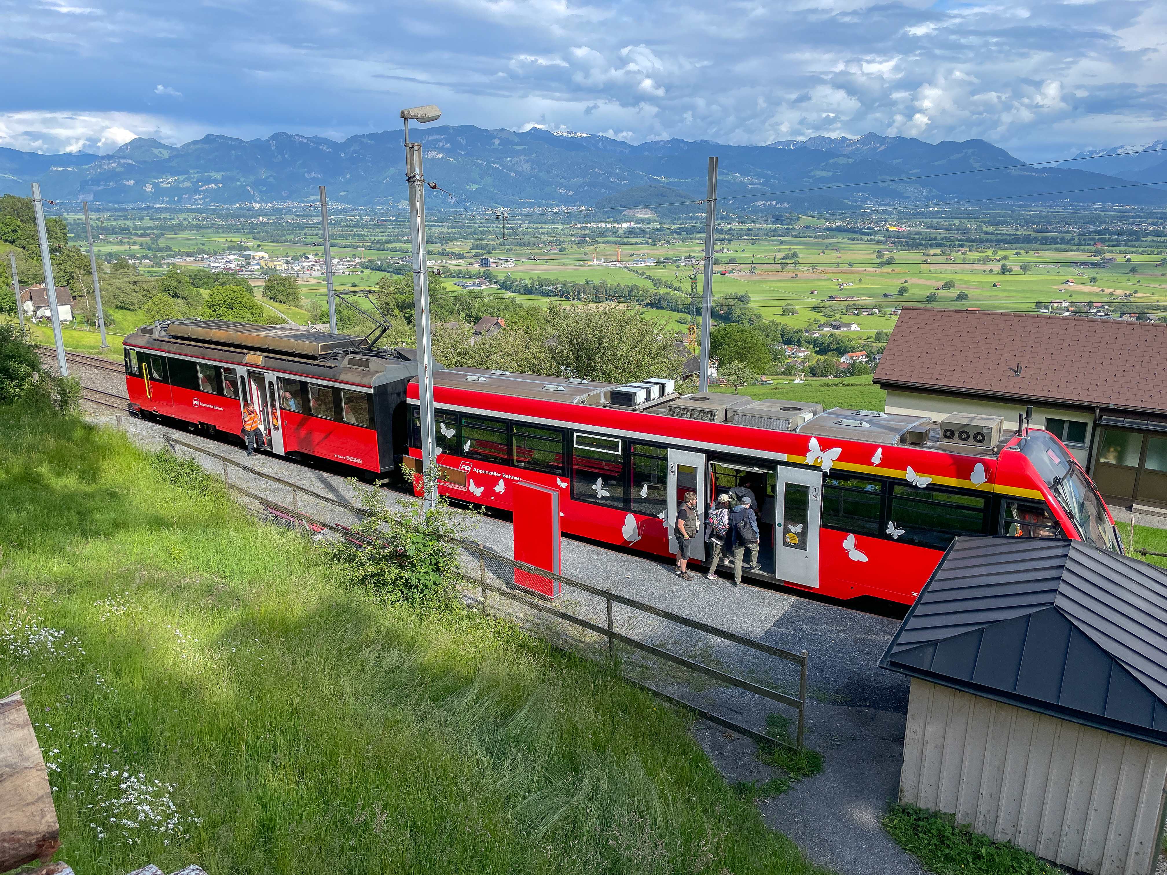 roter Zug an einer Haltestelle, im Hintergrund das St.Galler Rheintal