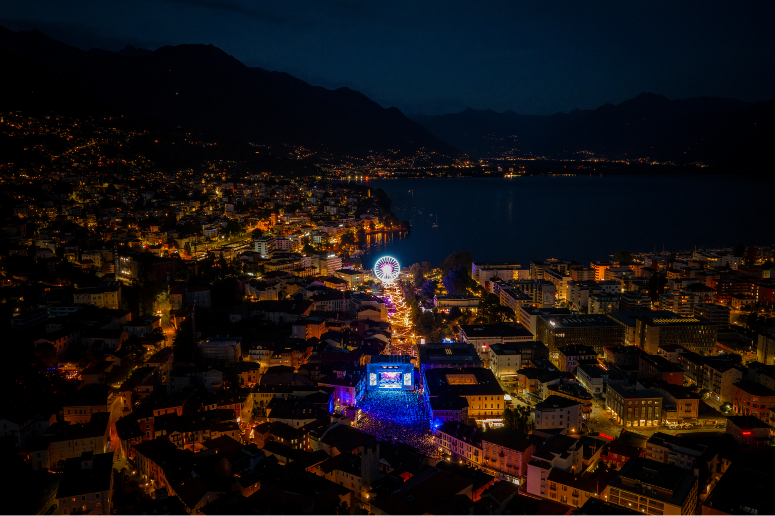 Vue aérienne nocturne de Locarno avec le site illuminé du festival Moon+Stars, la grande roue et le lac Majeur