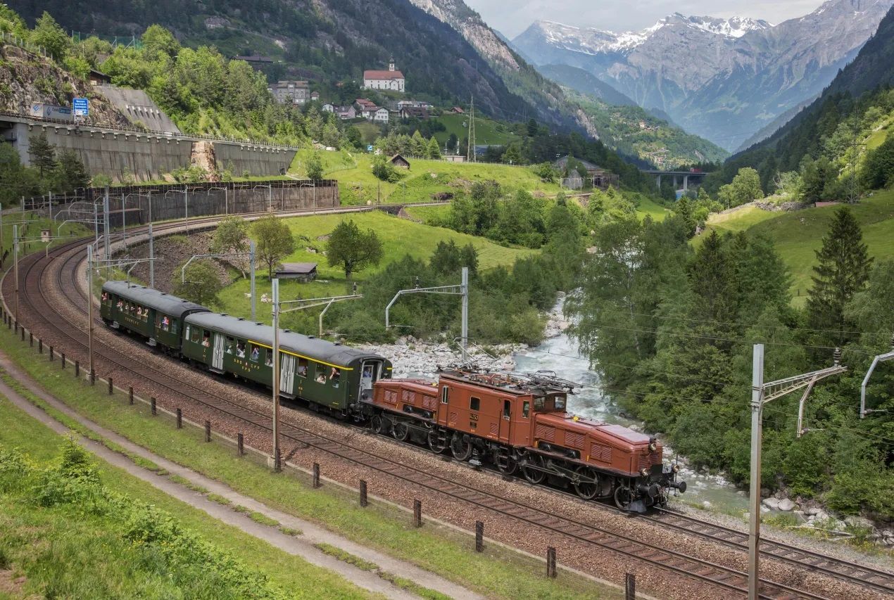 Treno storico della Ferrovia del Gottardo con locomotiva «Coccodrillo» sulla vecchia linea di montagna vicino a Wassen, circondato da prati verdi, un fiume e paesaggi alpini.