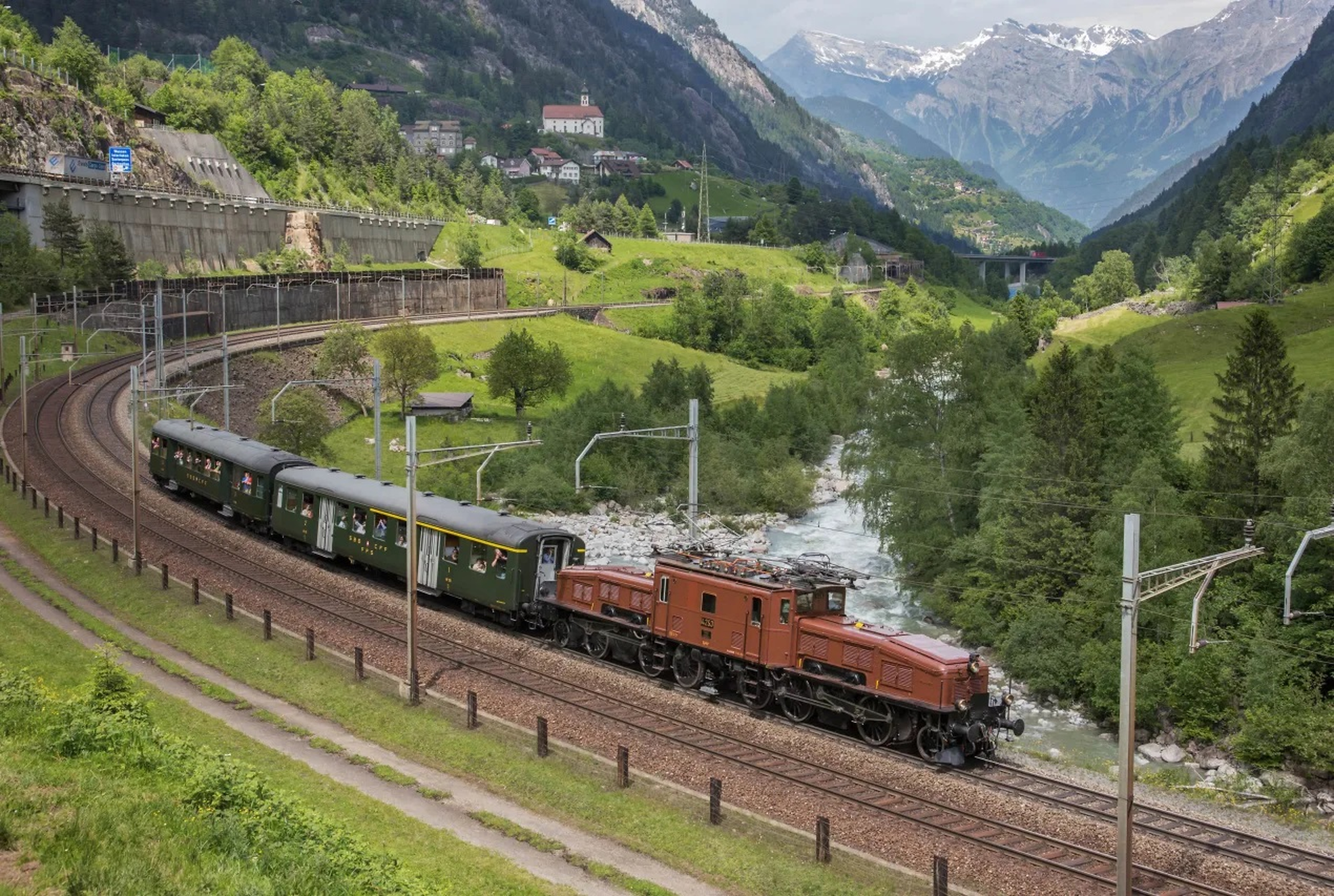 Historic Gotthard Railway train with Crocodile locomotive on the old mountain route near Wassen, surrounded by green meadows, a river, and Alpine scenery.
