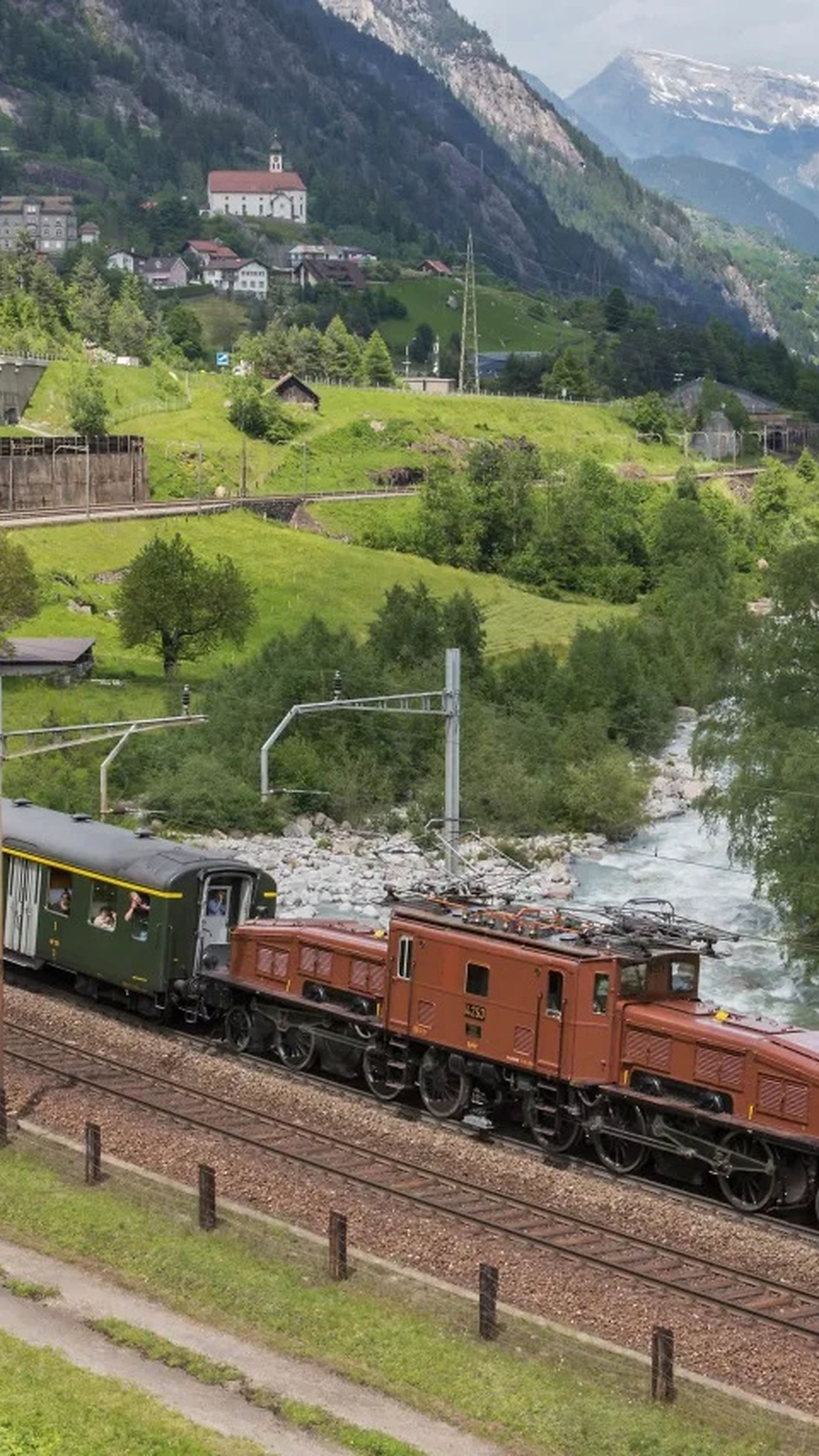 Historic Gotthard Railway train with Crocodile locomotive on the old mountain route near Wassen, surrounded by green meadows, a river, and Alpine scenery.