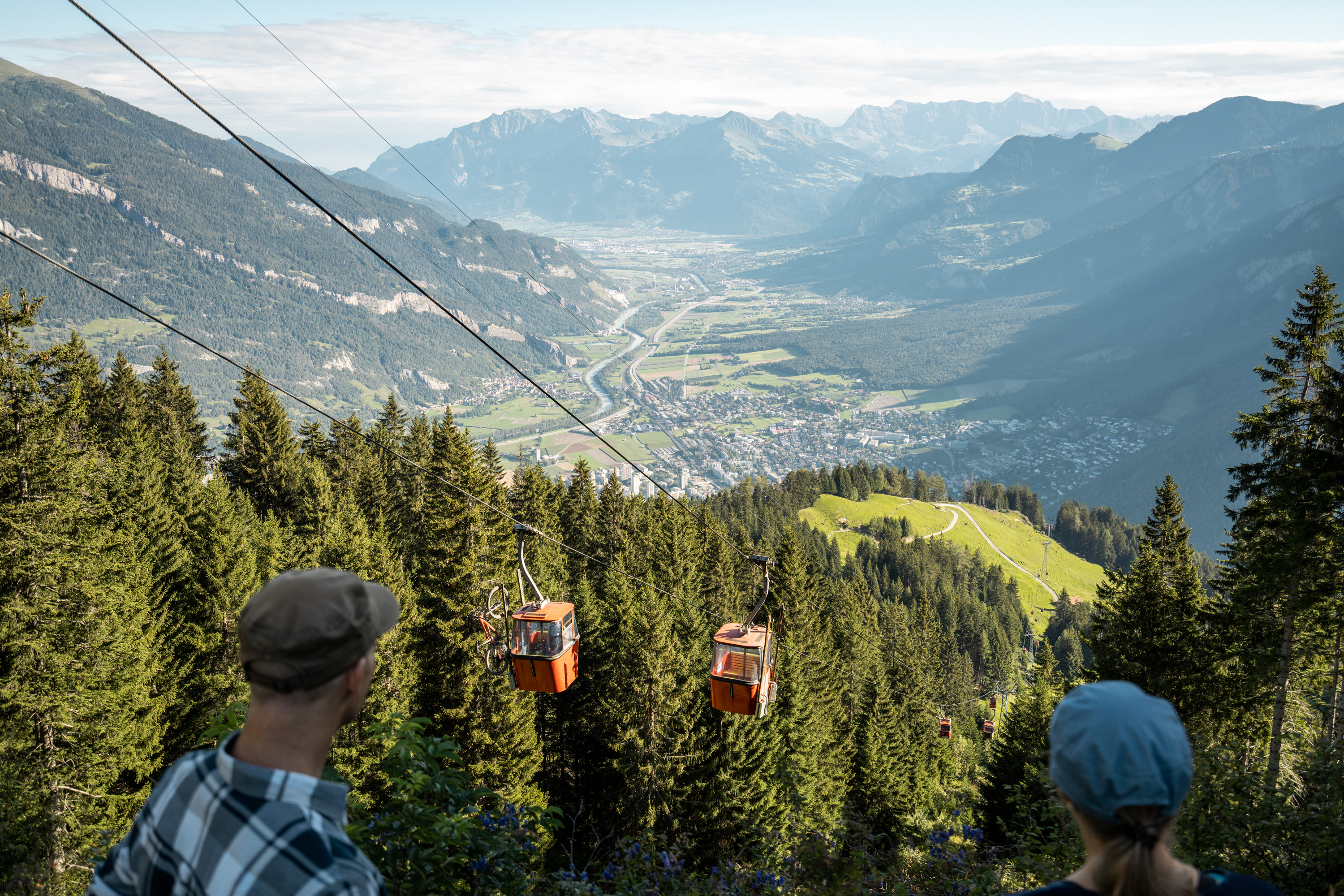 Two people looking at the red gondolas of the Brambrüesch cable car with a wide view over the Chur Rhine Valley.