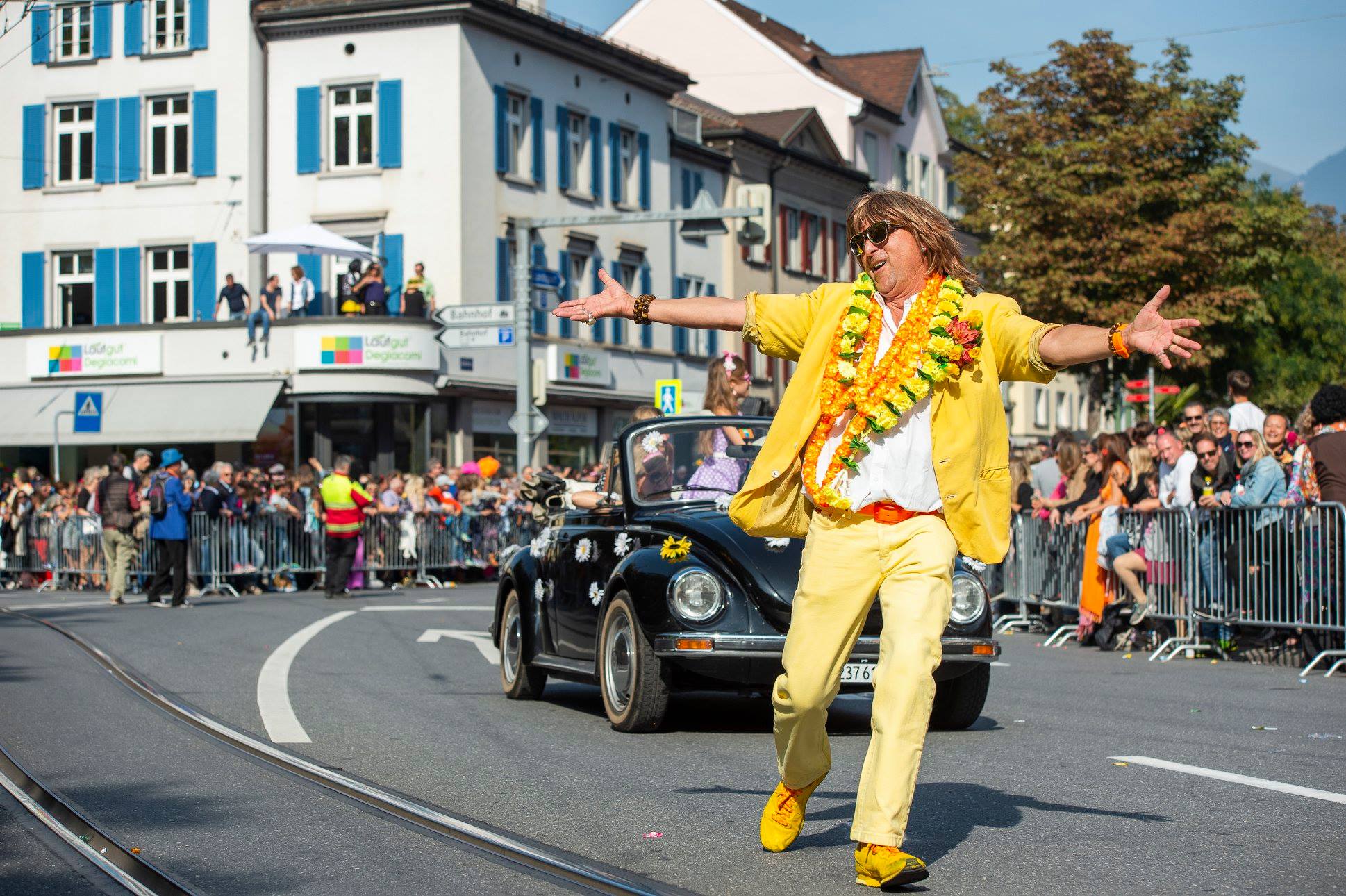 Die Teilnehmer der Churer Schlagerparade tanzen in gelben Retro-Outfits vor einem Oldtimer durch die festlich geschmückte Innenstadt, umringt von einer jubelnden Menschenmenge.