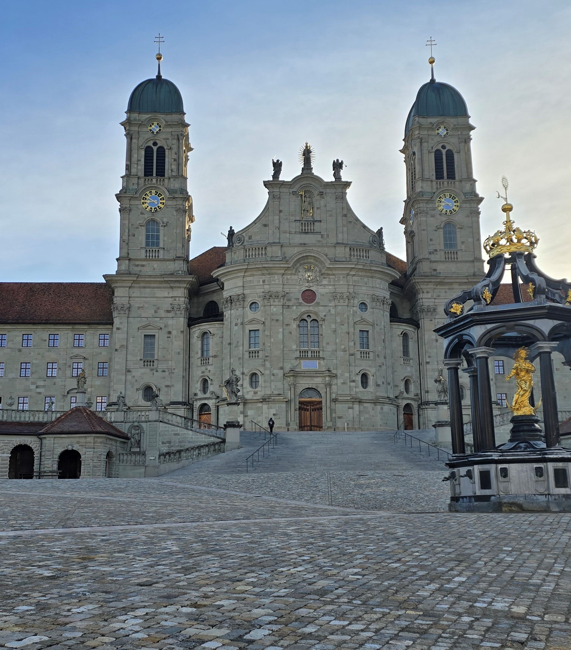Abbaye d’Einsiedeln avec façade baroque, ses deux tours et la fontaine mariale sur la place du monastère à Einsiedeln.