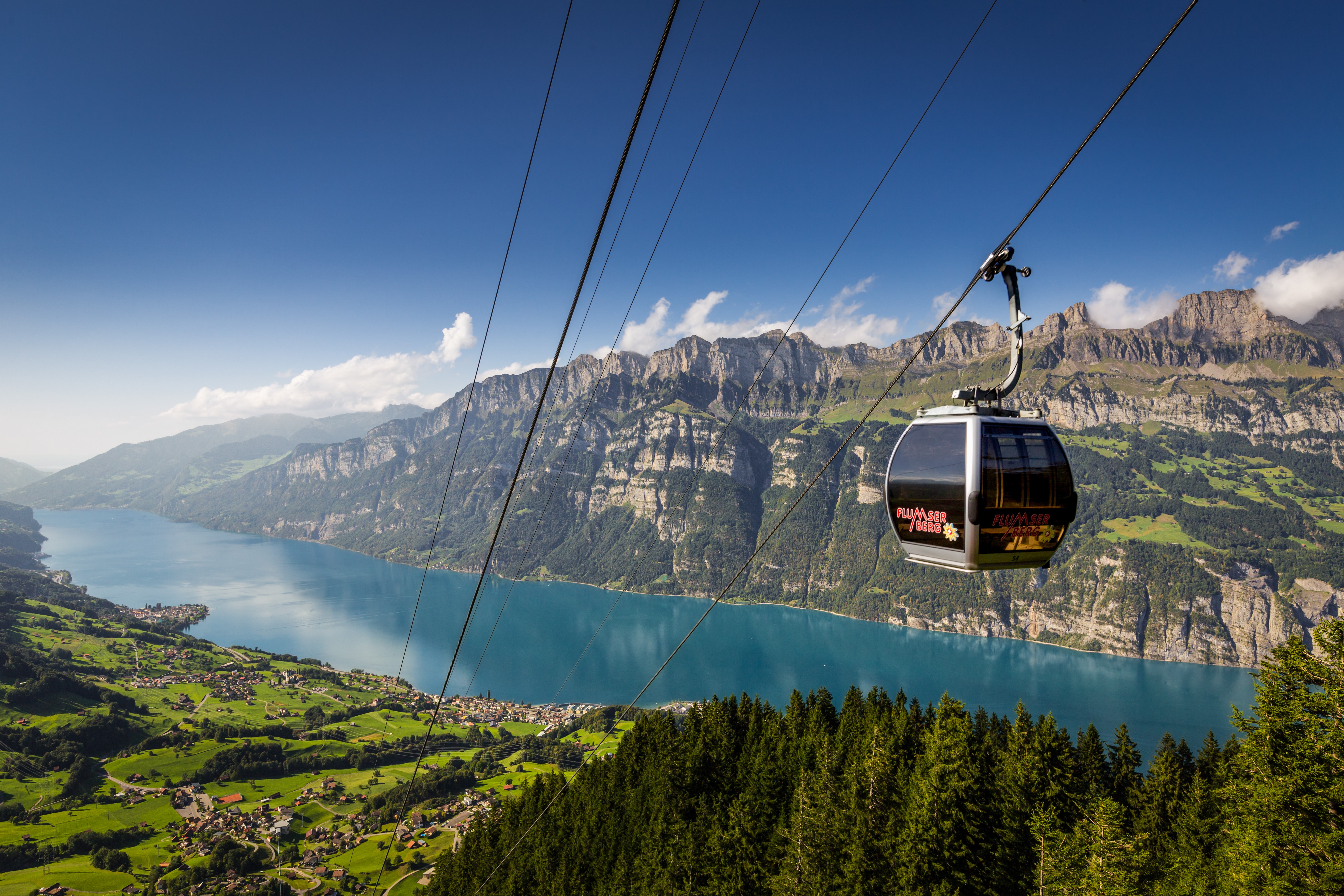 Une télécabine moderne du SeeJet survole les cimes d’arbres au Flumserberg – en arrière-plan, le lac de Walenstadt turquoise, des prairies verdoyantes et les falaises marquantes des Churfirsten